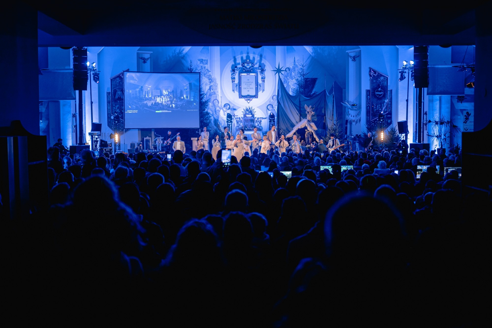 Zespół muzyczny na scenie podczas koncertu, oświetlony niebieskim światłem, widok zza publiczności filmującej występ telefonami.