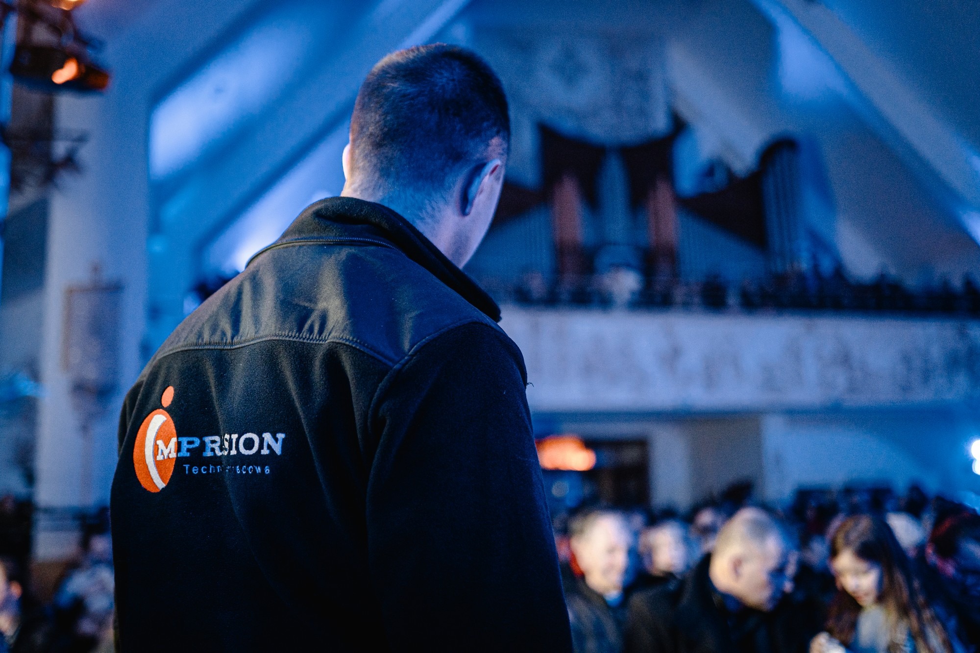 Osoba w czarnej bluzie z logo firmy MP RION, stojąca tyłem do kamery, obserwuje scenę z widowni podczas wydarzenia.
