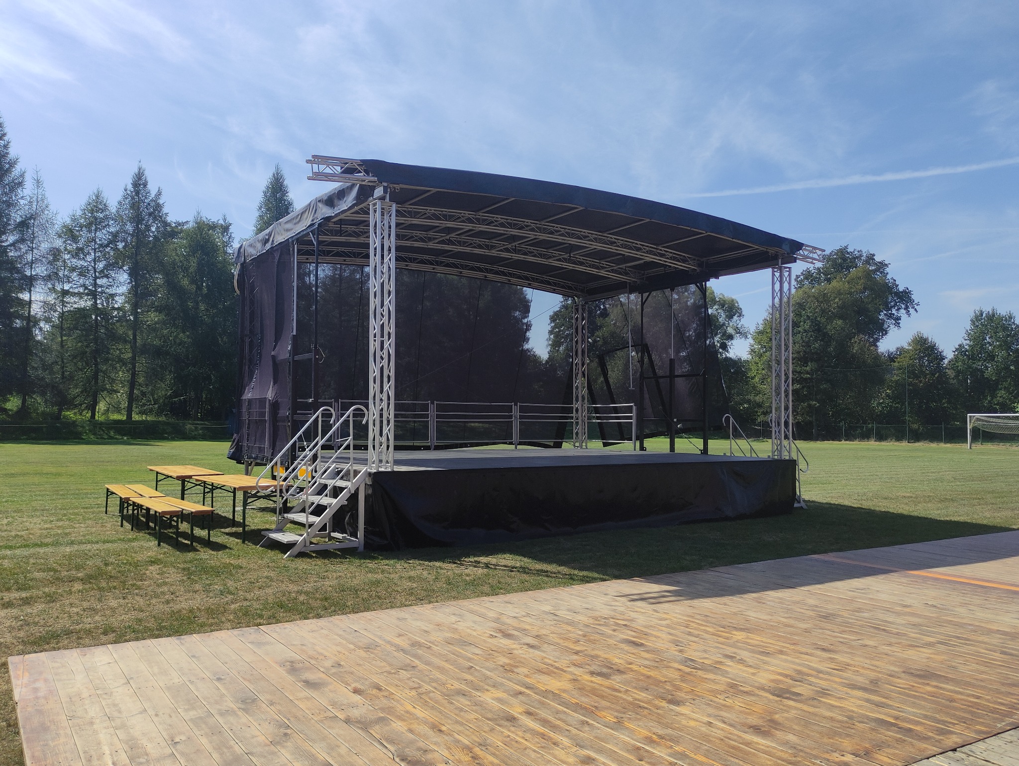 Plenerowa scena z metalową konstrukcją i czarnym materiałem, ustawiona na trawiastym terenie z drewnianym podestem z przodu i ławkami po lewej stronie, w tle drzewa i błękitne niebo.