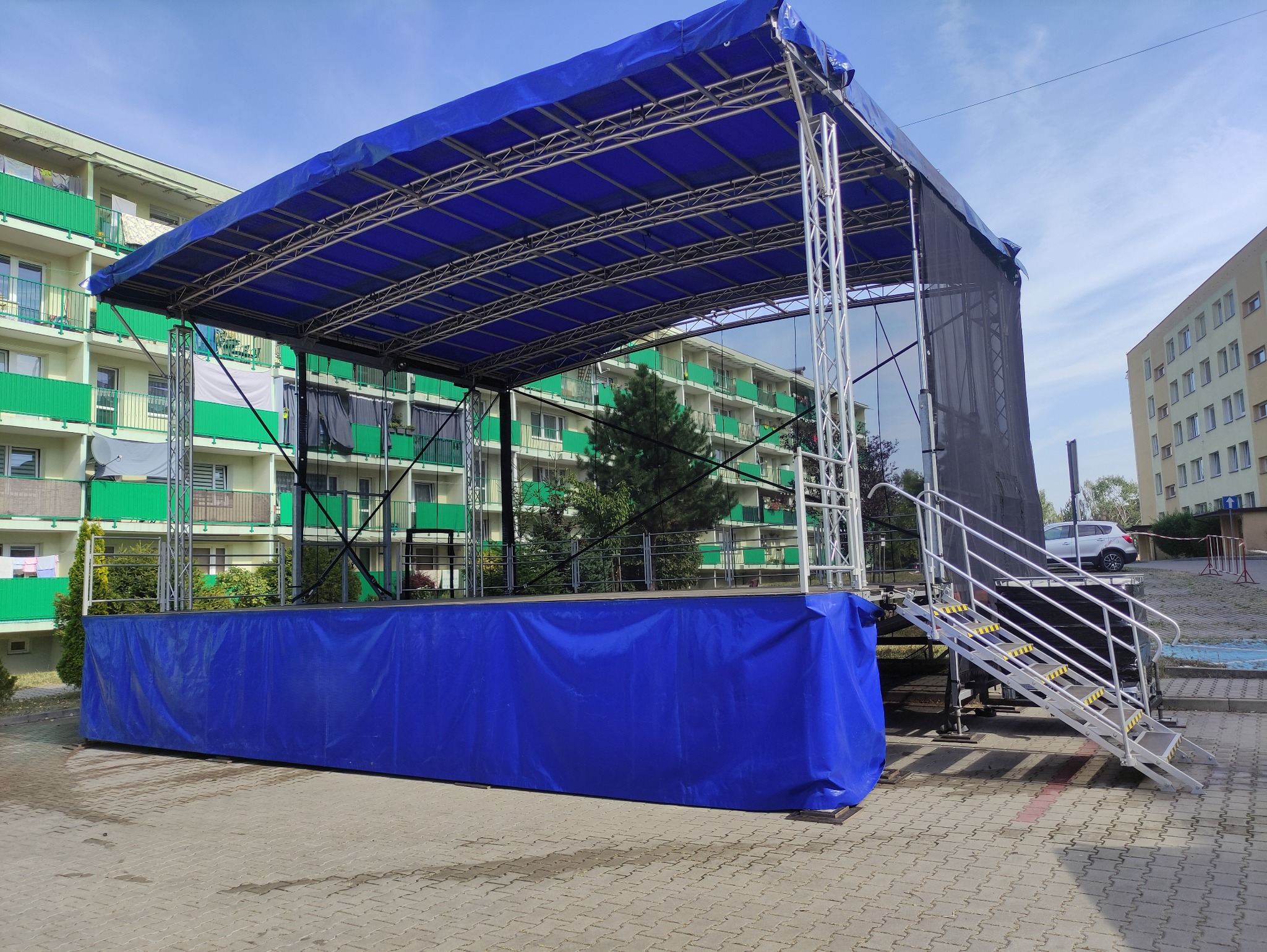 Rozłożona scena plenerowa z niebieskim zadaszeniem i srebrnymi schodami, na tle budynków mieszkalnych.