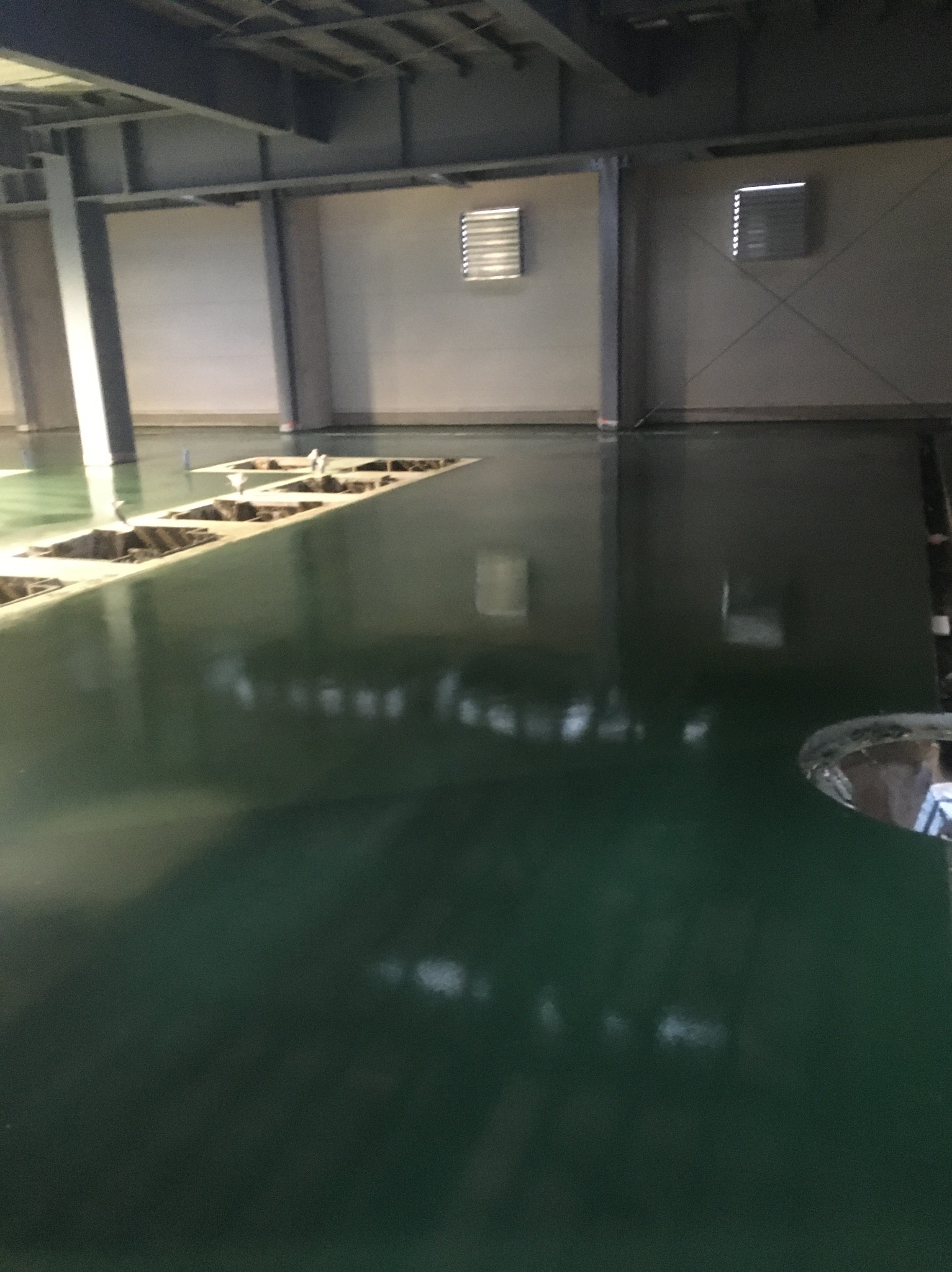 Wypoziomowana, błyszcząca, zielona podłoga w hali przemysłowej z otworami na instalacje, widoczne okratowane okna wentylacyjne w tle.