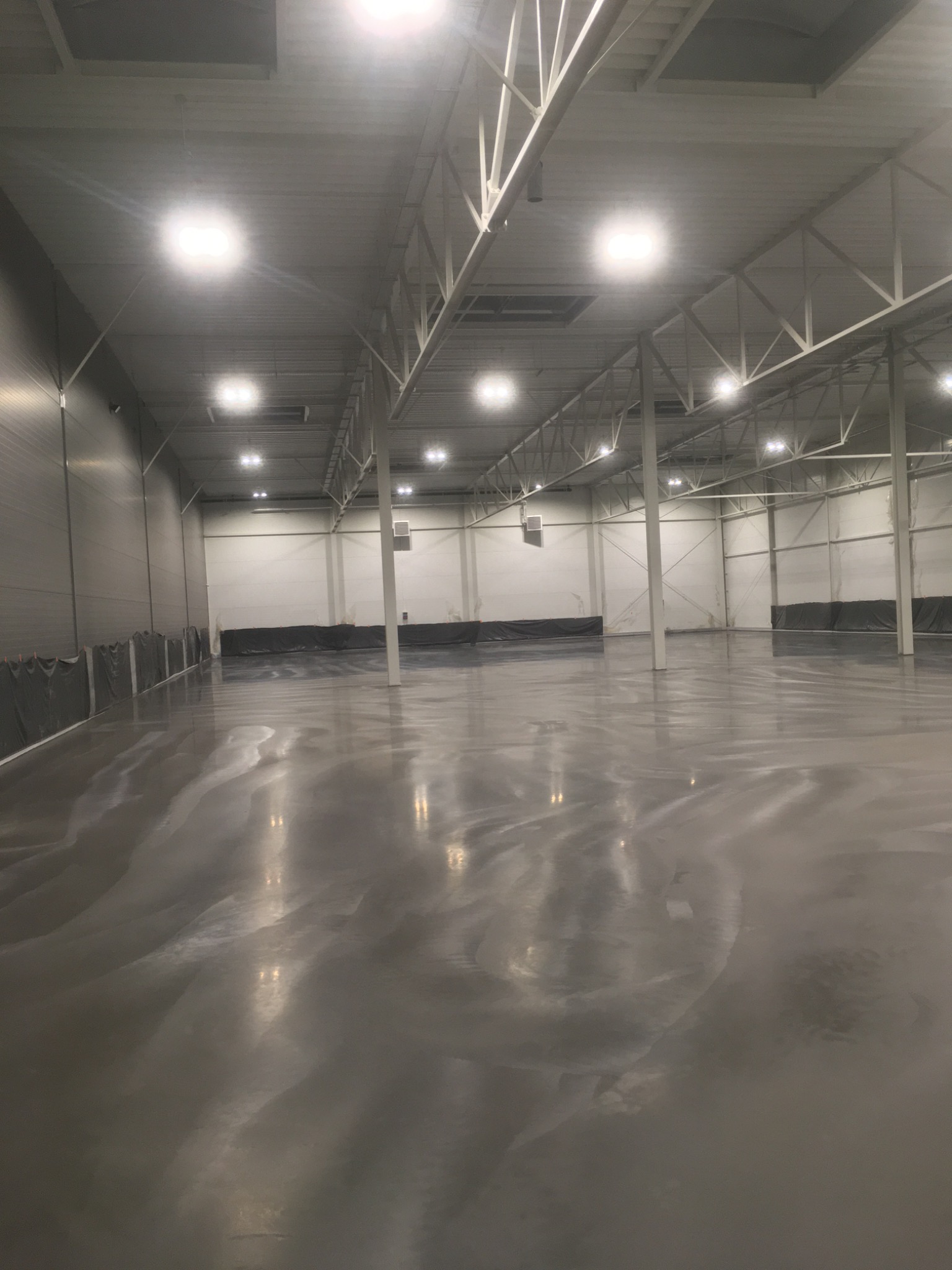 Błyszcząca, szara posadzka w pustym, oświetlonym hali magazynowej z widoczną metalową konstrukcją dachu.