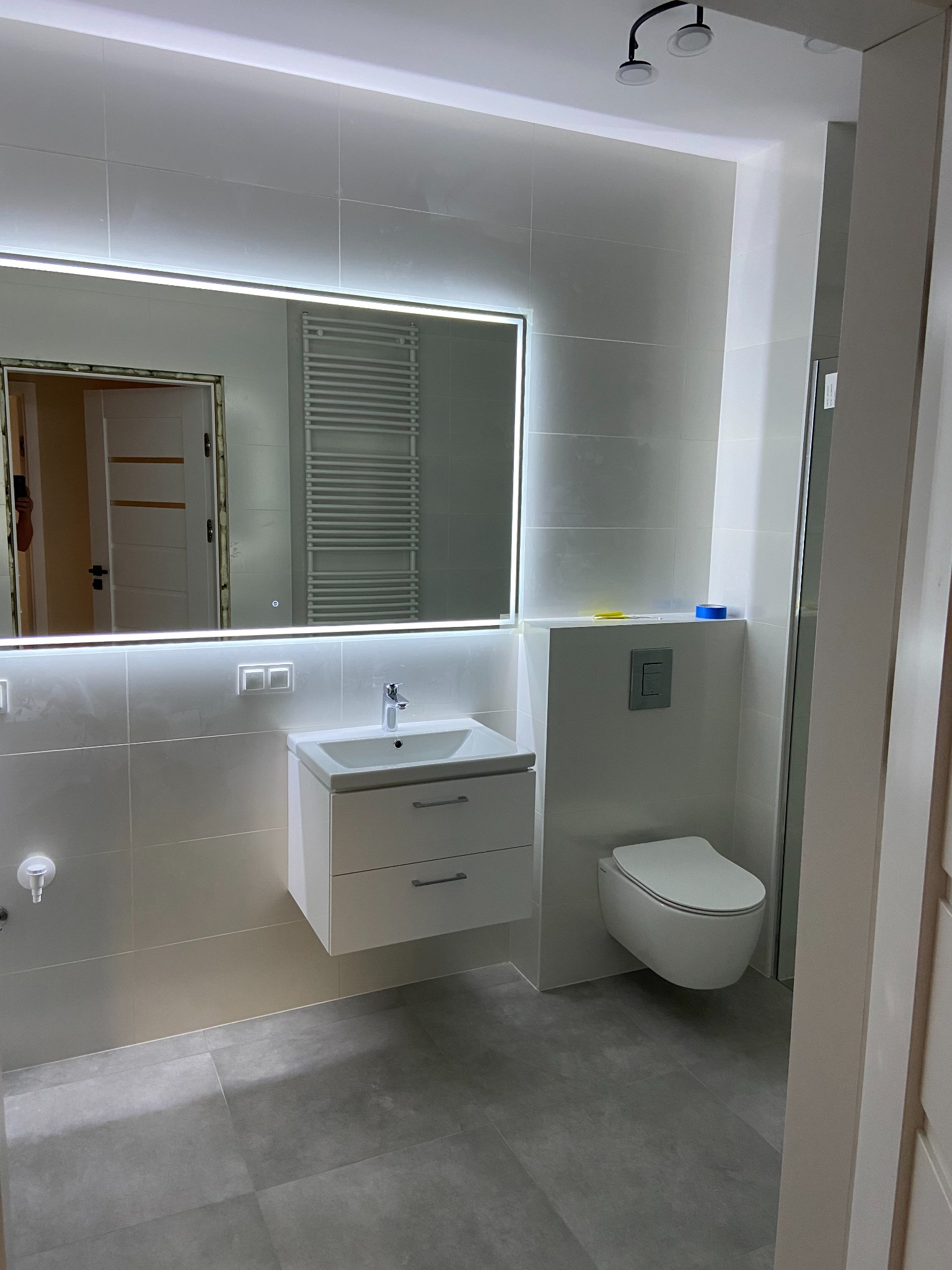 Nowoczesna łazienka z podświetlanym lustrem, wiszącą toaletą i umywalką z szafką, wykończona szarymi płytkami. W odbiciu lustra widoczny grzejnik drabinkowy.