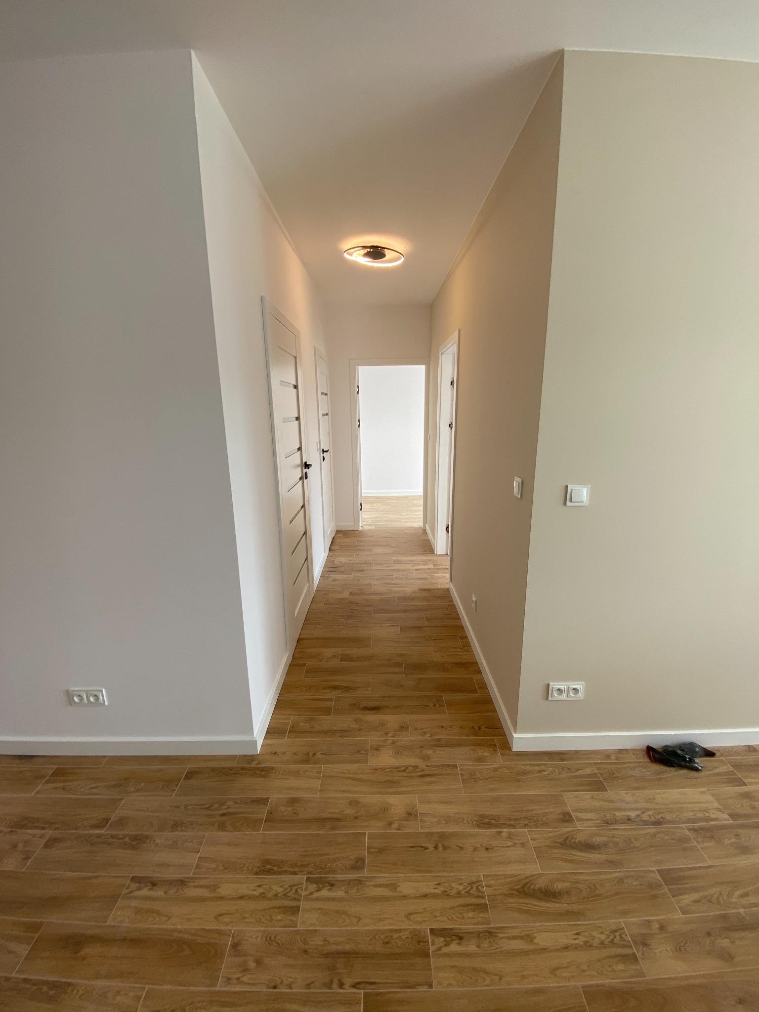 Długi korytarz w jasnych barwach z drewnopodobną podłogą, widoczne białe ściany, drzwi z czarnymi klamkami i gniazdka elektryczne przy podłodze. Na podłodze leżą czarne rękawice.