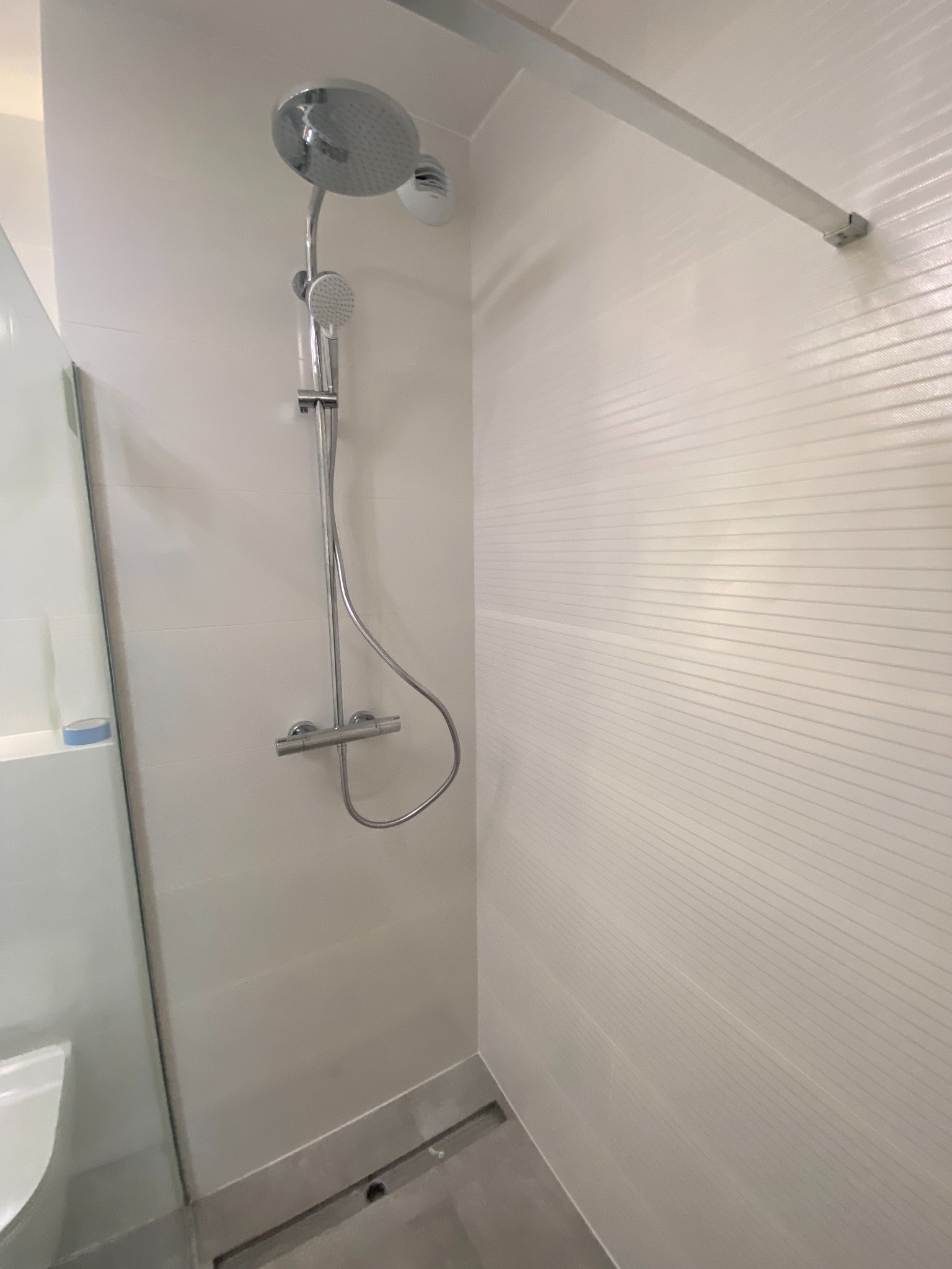 Nowoczesna kabina prysznicowa z deszczownicą i baterią termostatyczną, wykończona białymi płytkami z subtelnym poziomym wzorem, szklane drzwi kabiny z widoczną krawędzią.