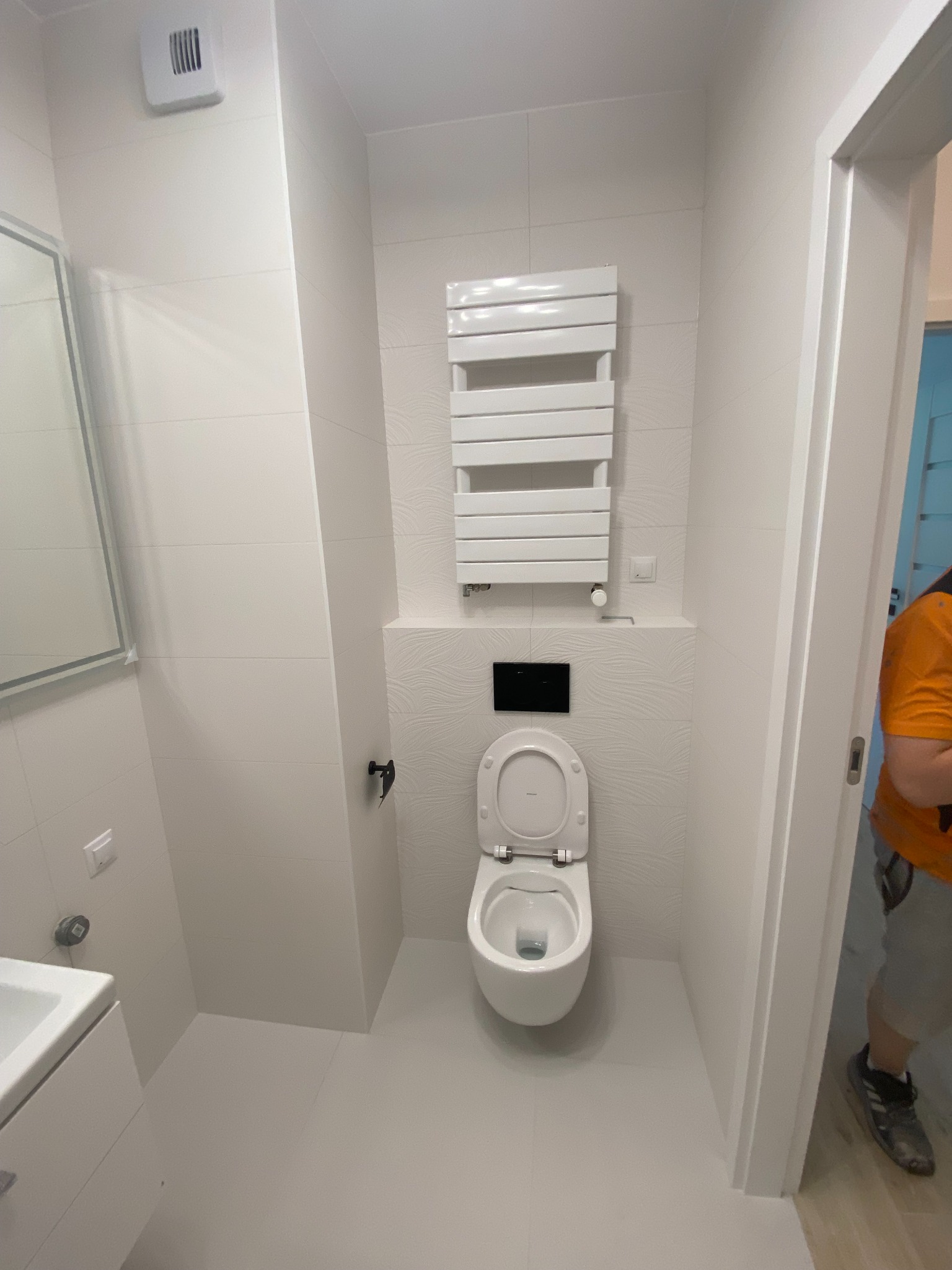 Jasna, wykafelkowana łazienka z wiszącą toaletą z otwartą klapą, grzejnikiem drabinkowym i elementami armatury w kolorze czarnym, widziana z perspektywy drzwi z fragmentem osoby w tle.