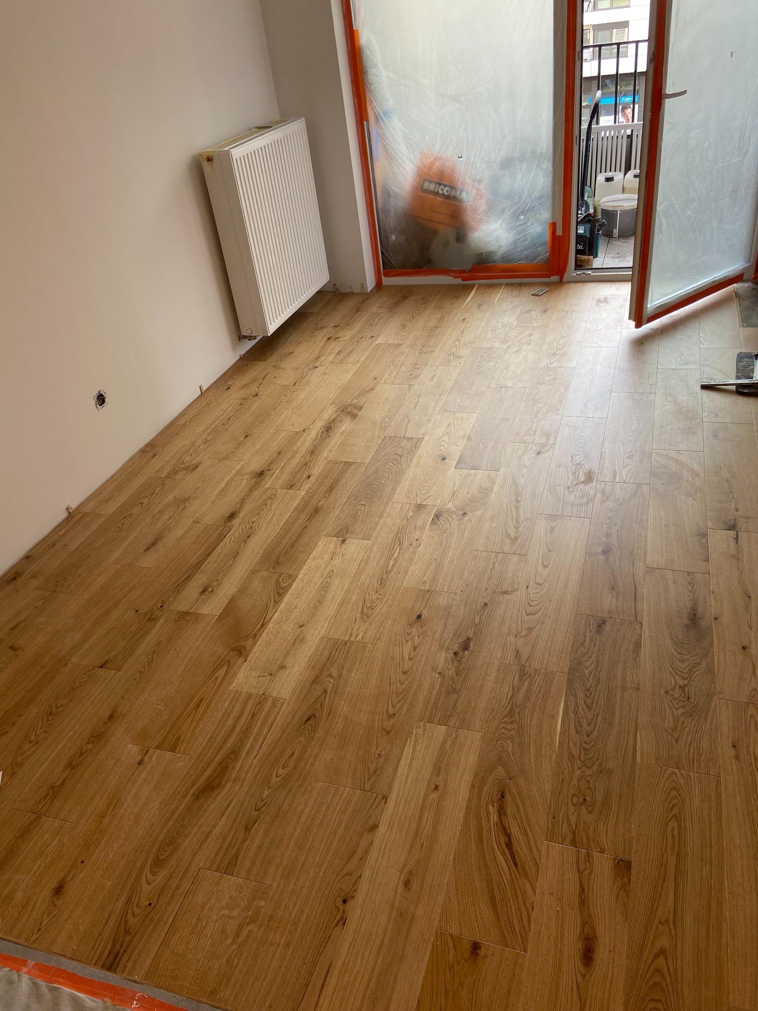 Nowo ułożone, jasne panele podłogowe z widocznymi szczelinami dylatacyjnymi przy ścianie, zabezpieczone klinami, z grzejnikiem na ścianie i folią ochronną na drzwiach balkonowych.