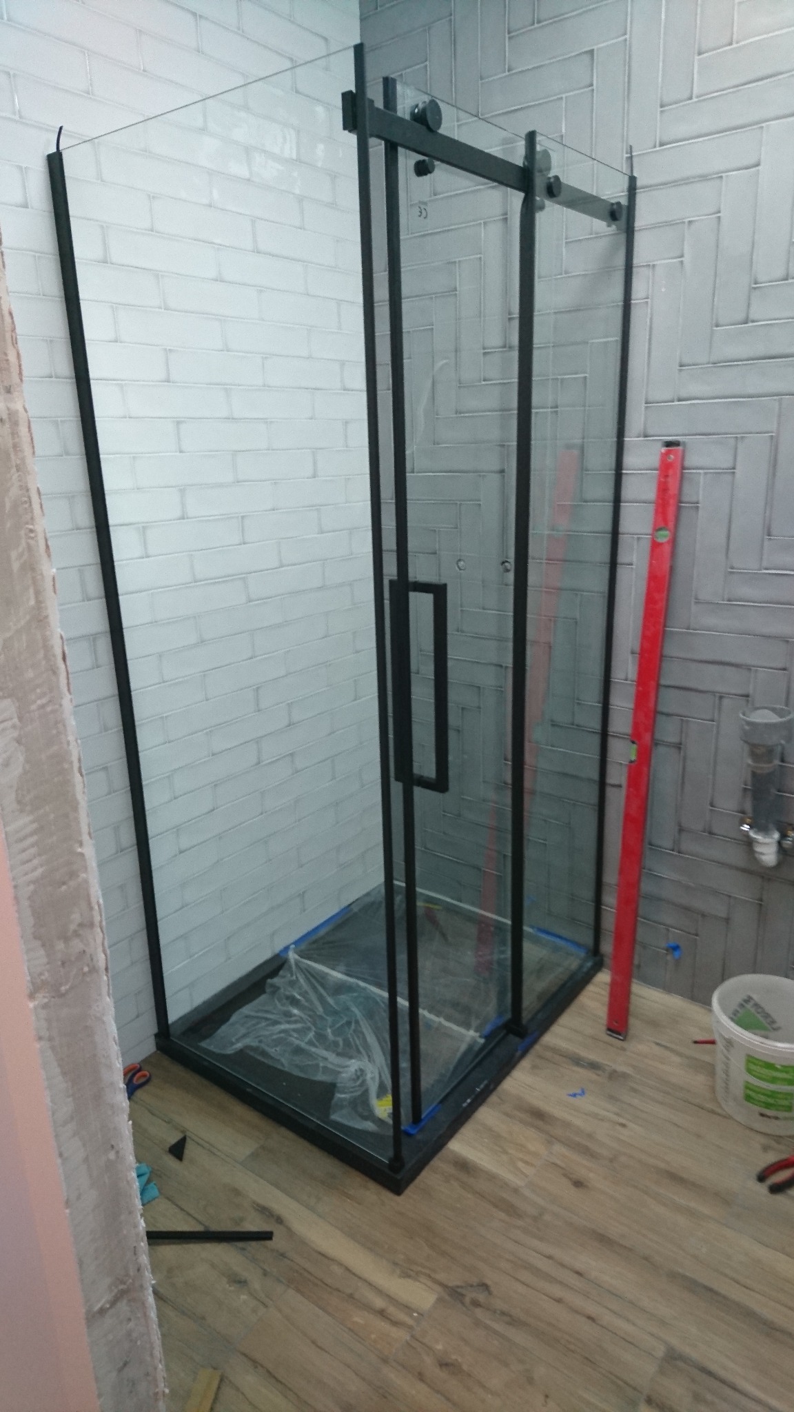 Montaż kabiny prysznicowej z czarnymi profilami na tle szarej cegły, widoczna poziomica i folia ochronna na brodziku.