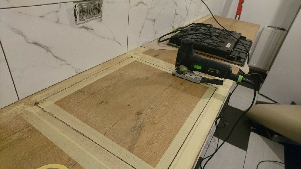 W Warszawie, wyrzynarka Festool wycina otwór w drewnianym blacie, obklejonym taśmą malarską, pod płytę indukcyjną; widoczne płytki na ścianie.