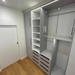 Brzostowski Meble - Szara szafa z oświetleniem LED, z szufladami z czarnymi uchwytami i półkami, widok na wnętrze szafy z drążkiem na ubrania i półką, drewniana podłoga, białe drzwi obok.