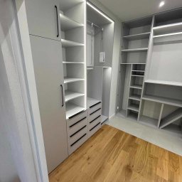 Brzostowski Meble - Jasnoszara garderoba z oświetleniem LED wewnątrz, z czarnymi, minimalistycznymi uchwytami na frontach szuflad i drzwi, na parkiecie z naturalnego drewna.