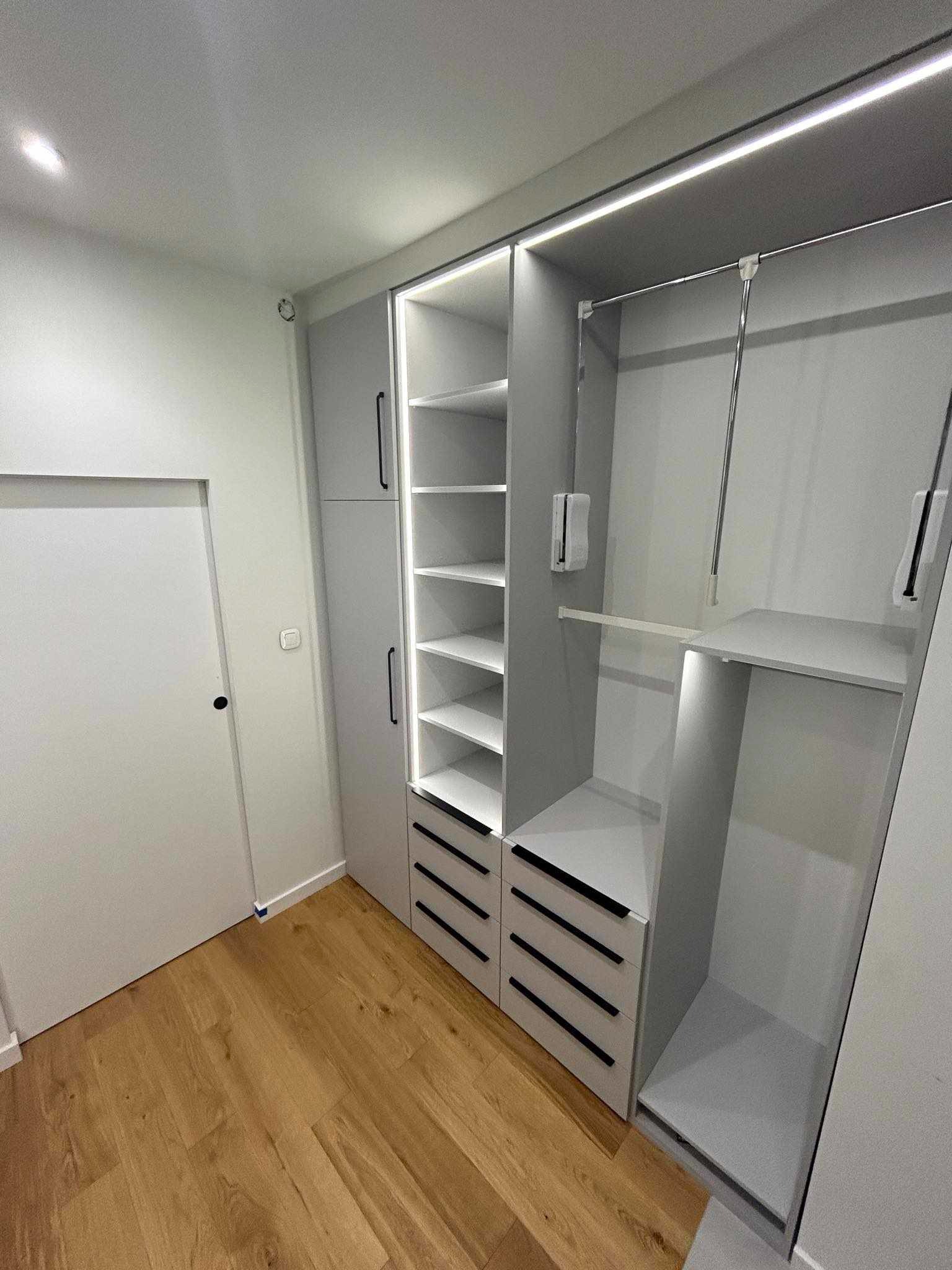 Szara szafa z oświetleniem LED, z szufladami z czarnymi uchwytami i półkami, widok na wnętrze szafy z drążkiem na ubrania i półką, drewniana podłoga, białe drzwi obok.