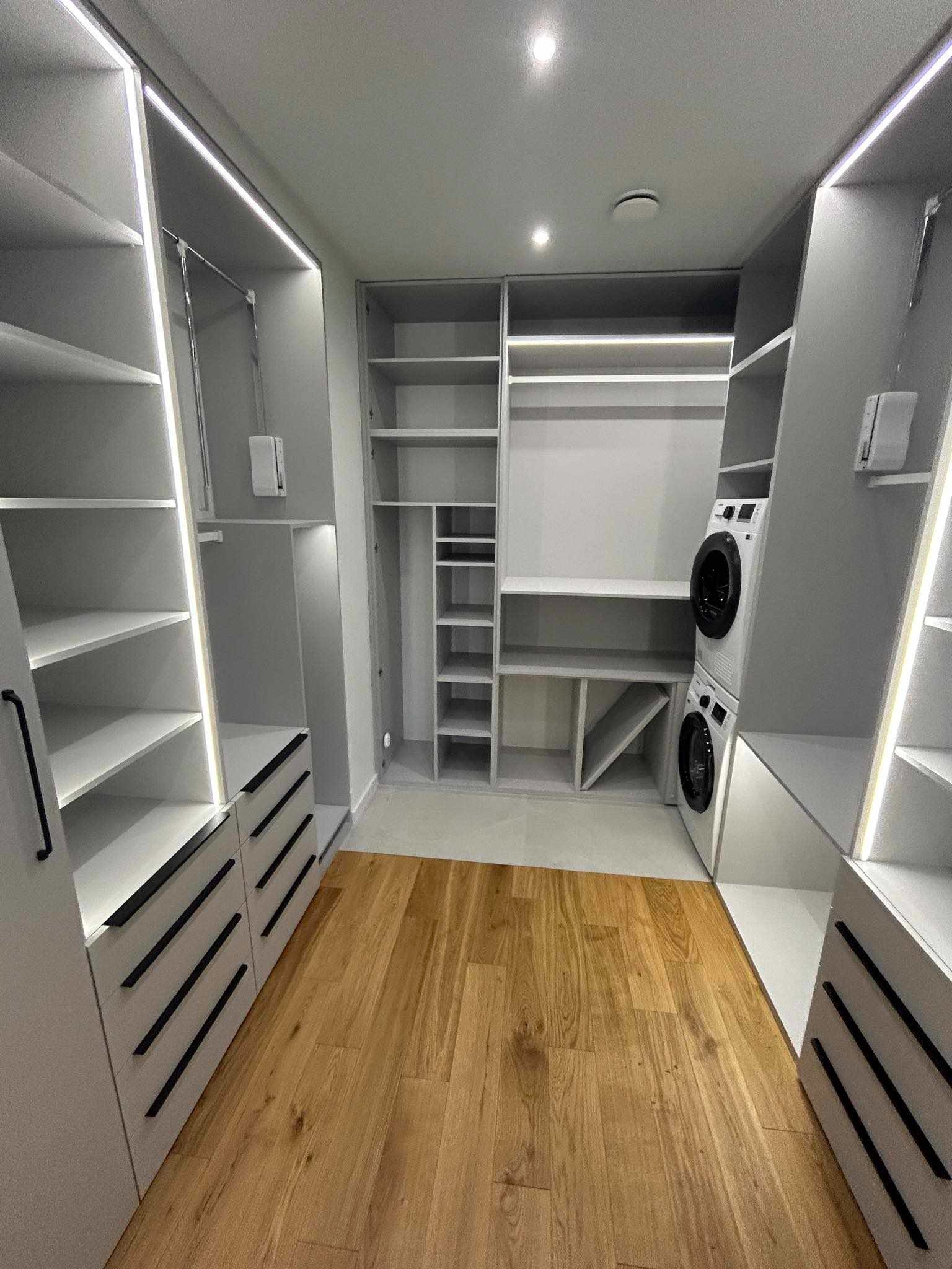 Jasnoszara garderoba na wymiar z oświetleniem LED, drewnianą podłogą i pralką w zabudowie.