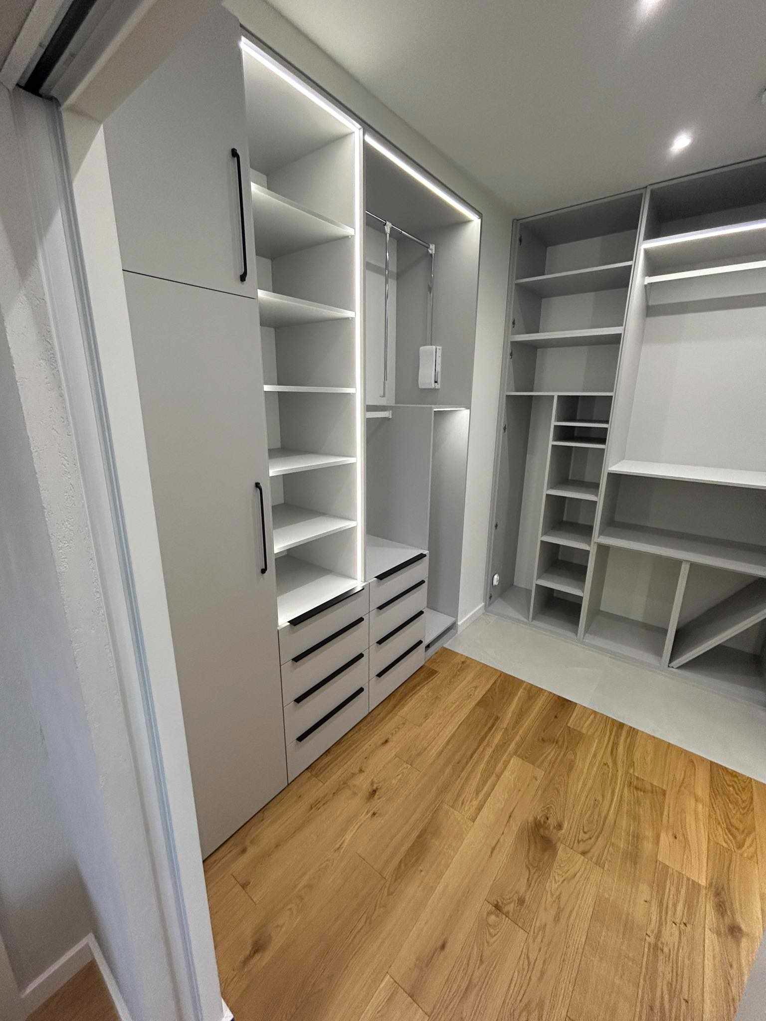 Jasnoszara garderoba z oświetleniem LED wewnątrz, z czarnymi, minimalistycznymi uchwytami na frontach szuflad i drzwi, na parkiecie z naturalnego drewna.