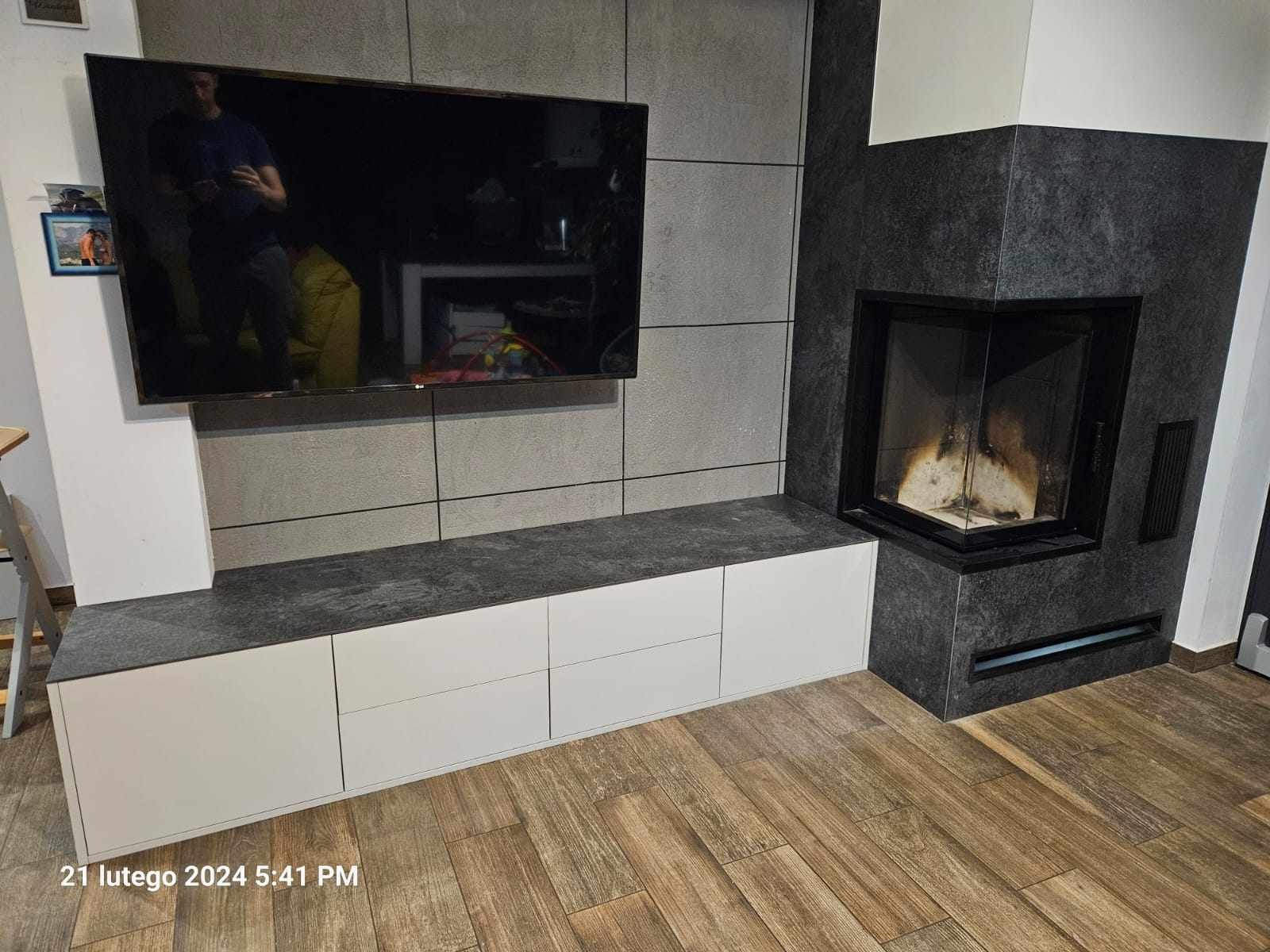 Nowoczesna zabudowa ściany z szarych paneli, białej szafki RTV z szarym blatem i narożnego kominka z czarnego kamienia, na podłodze panele imitujące drewno.