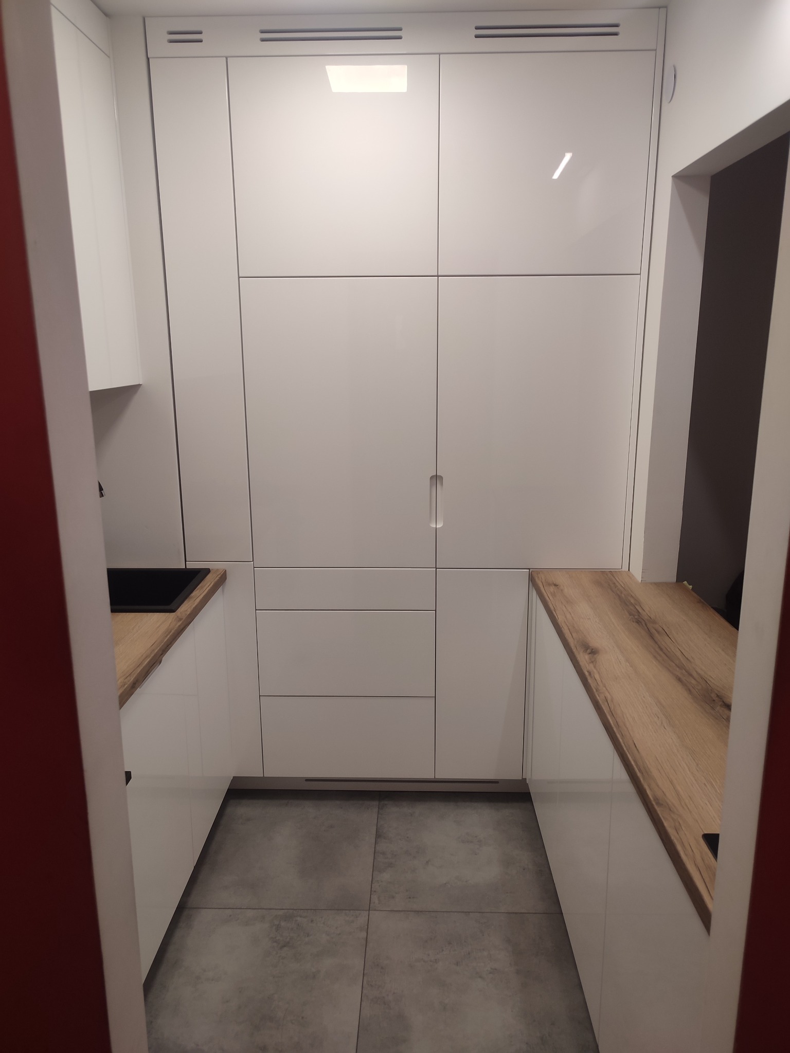 Wnętrze małej, nowoczesnej kuchni w Warszawie z zabudową meblową w kolorze białym i drewnianym blatem, widok z perspektywy wejścia.