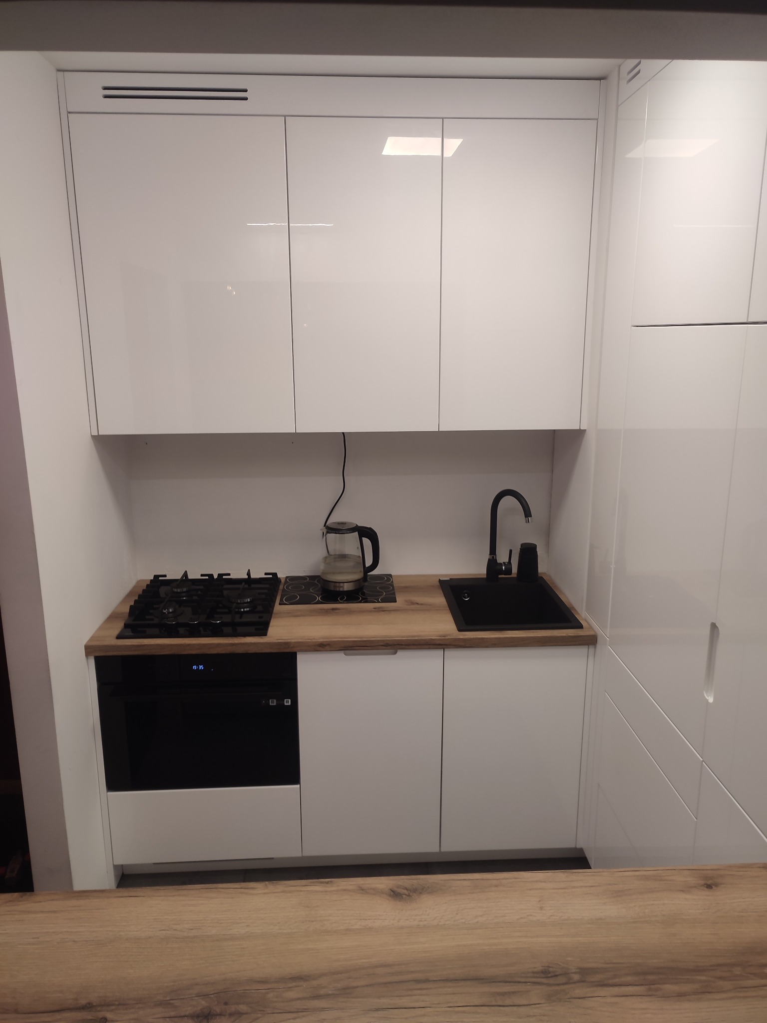 Biała, minimalistyczna kuchnia z czarnym zlewem, płytą grzewczą i piekarnikiem, oświetlona naturalnym światłem, z czajnikiem na blacie.