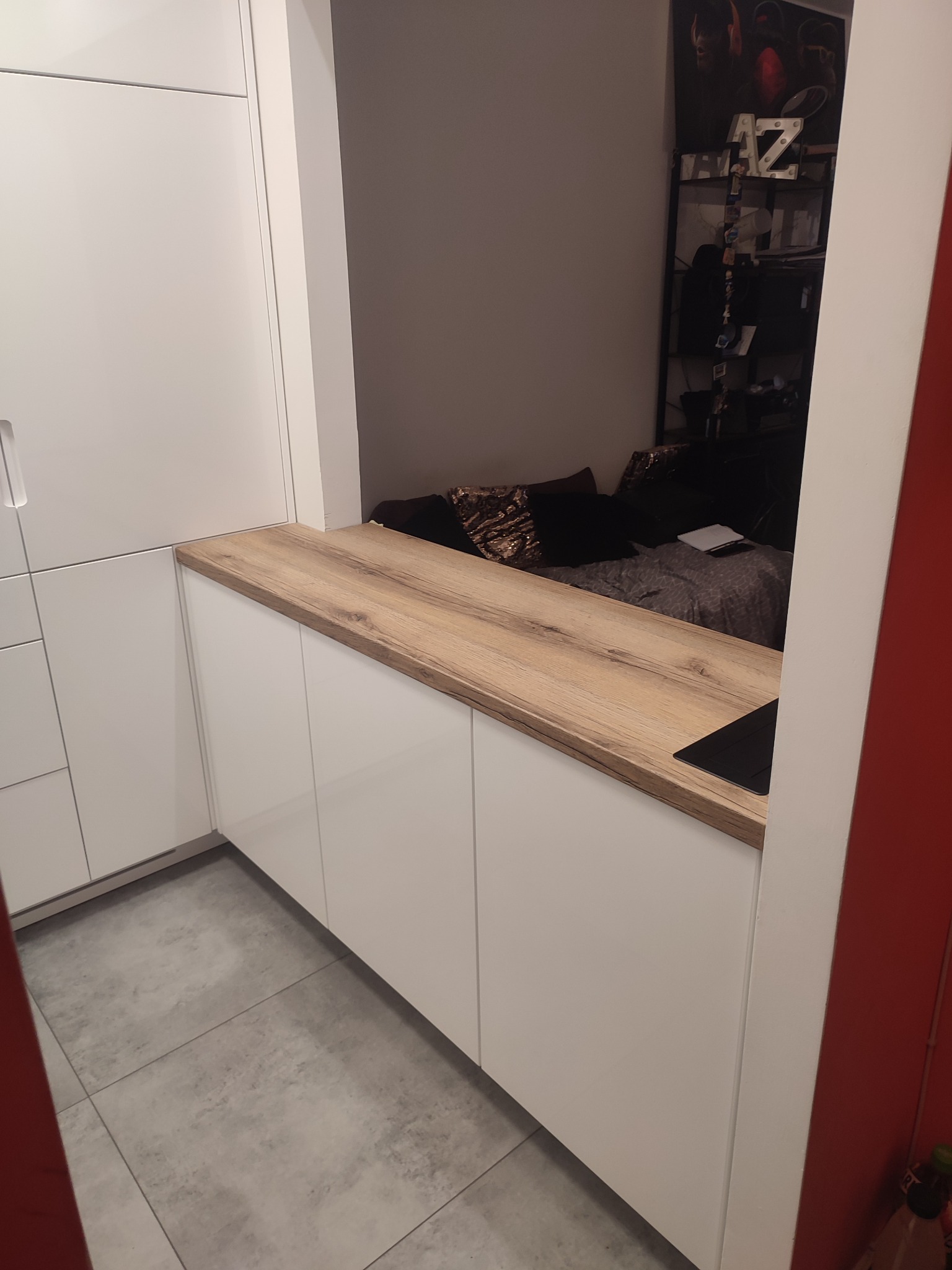 Biała zabudowa kuchenna z drewnianym blatem przechodząca w ściankę działową, za którą widać fragment pokoju z półką i dekoracjami.