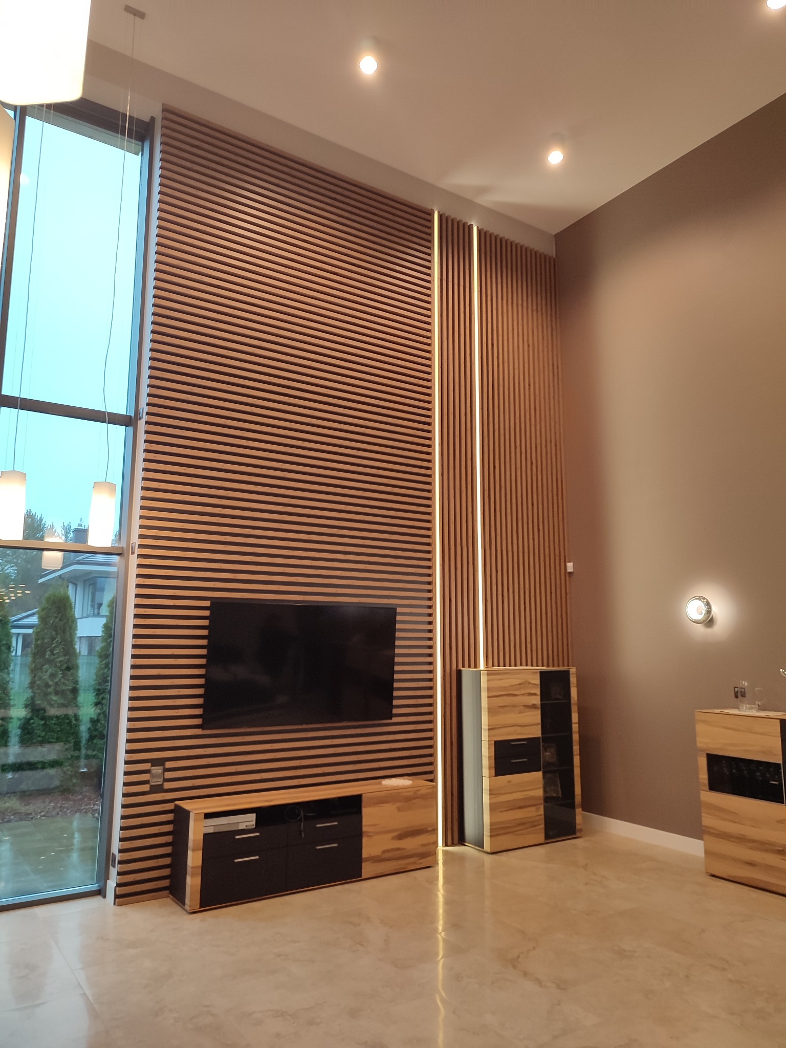 Nowoczesny salon z drewnianą ścianą z listew, podświetleniem LED, telewizorem i szafkami RTV z forniru w odcieniach brązu i czerni.