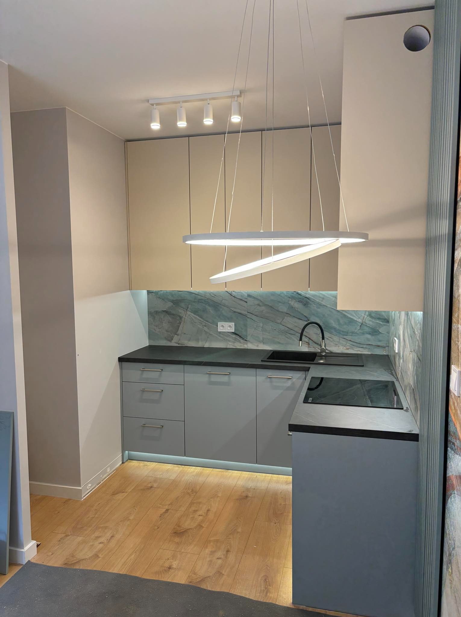 Nowoczesna kuchnia z szarymi szafkami, czarnym blatem i marmurowym backsplashiem, oświetlona designerską lampą wiszącą i listwą LED pod szafkami.