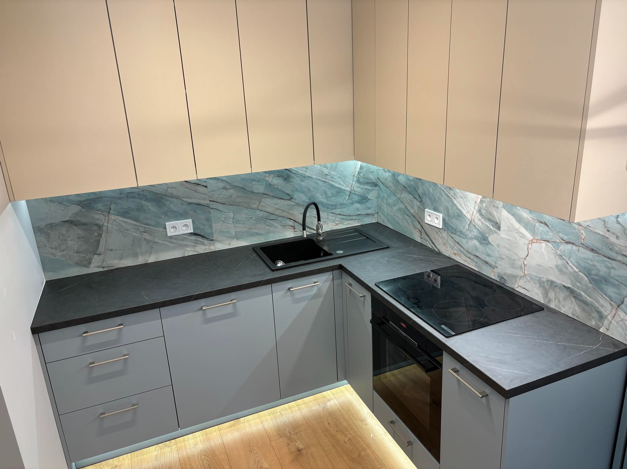Nowoczesna kuchnia z szarymi szafkami, blatem z ciemnego kamienia i marmurowym panelem ściennym, z widoczną płytą indukcyjną i oświetleniem pod szafkami.