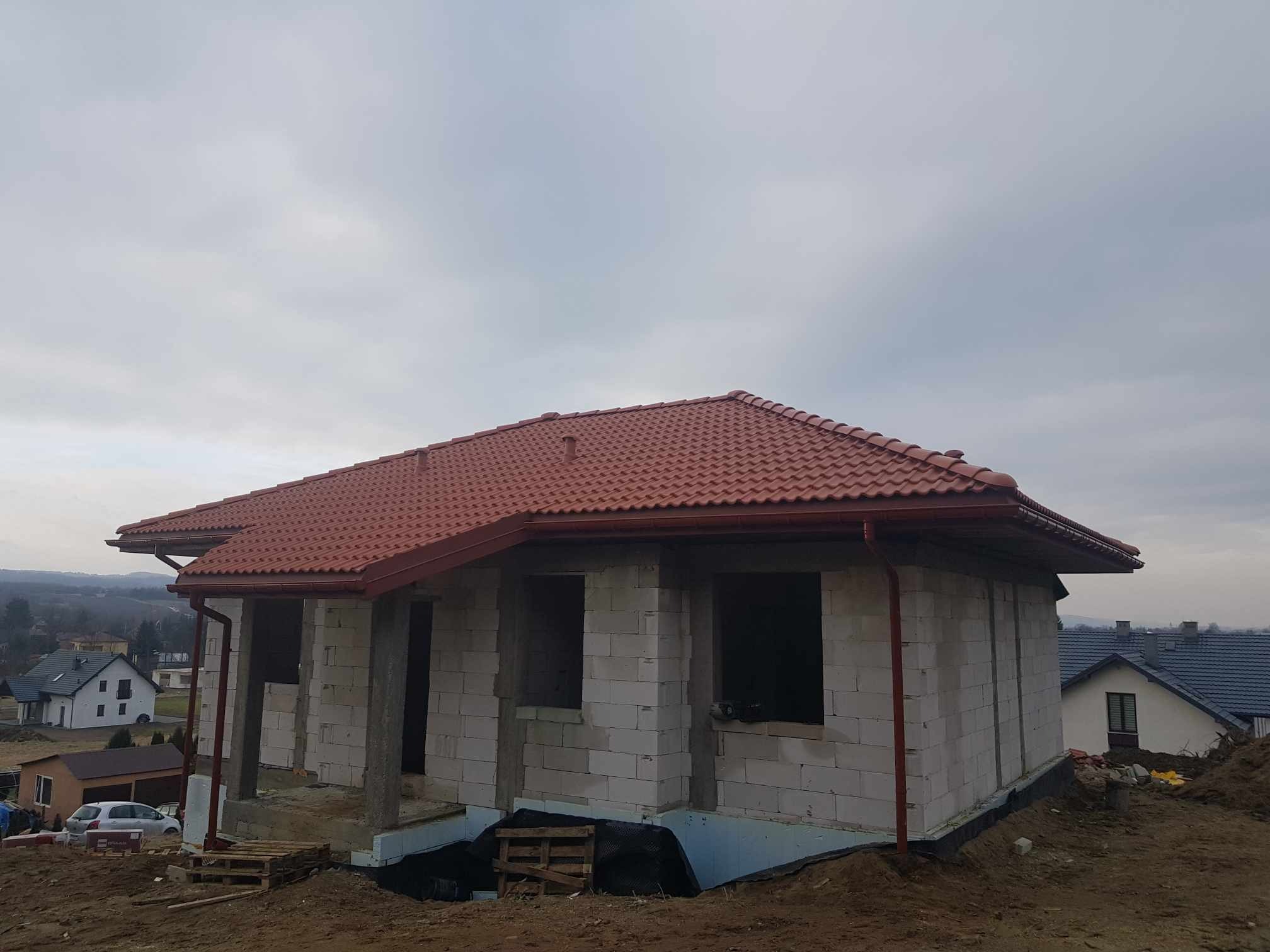 Dom w trakcie budowy z czerwoną dachówką ceramiczną i zamontowanymi rynnami, widoczne bloczki betonowe i przygotowany teren.