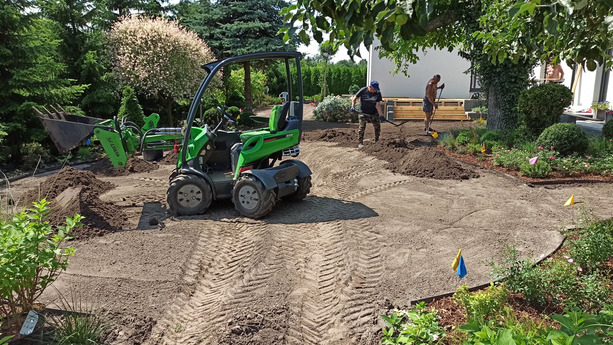 Przygotowanie terenu pod ogród: zielony miniładowarka Avant i dwóch mężczyzn wyrównujących ziemię podłoże, widoczne ślady opon na piasku, w tle drzewa i budynek mieszkalny.
