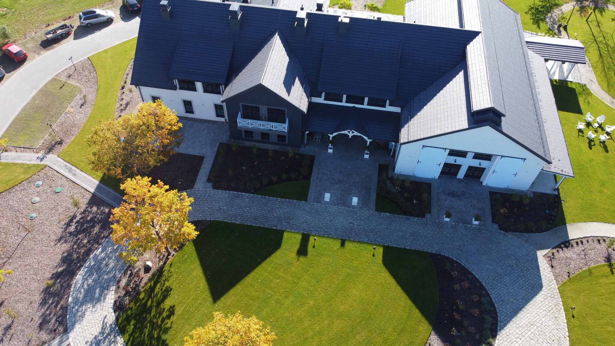 Widok z lotu ptaka na duży dom z ciemnym dachem, otoczony zadbanym trawnikiem, ścieżkami z kostki brukowej i drzewami o jesiennych liściach. W tle widoczne zaparkowane samochody. Na trawniku...