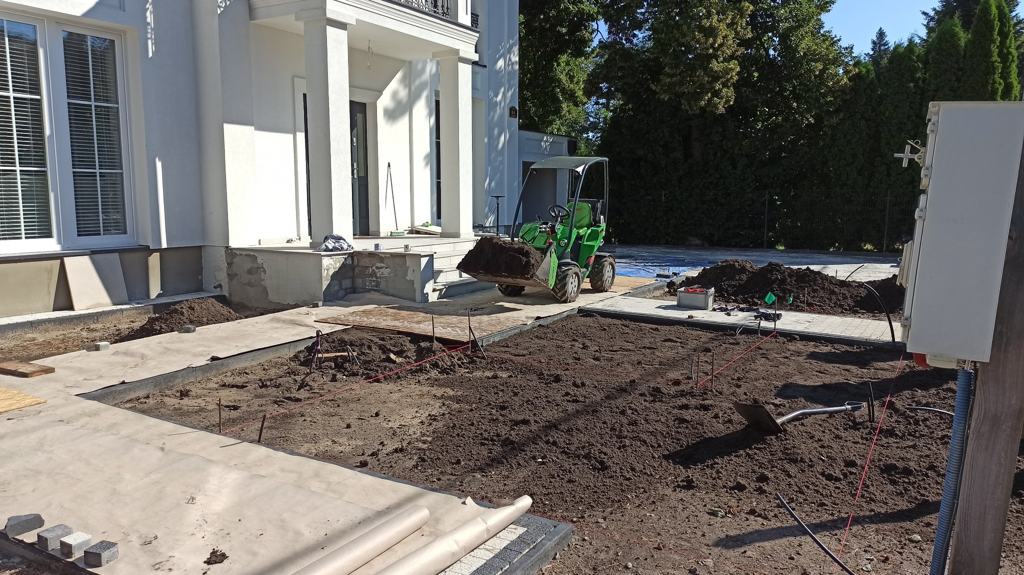 Przygotowanie terenu pod trawnik: miniładowarka z pełną łyżką ziemi, wytyczone rabaty z rozciągniętymi sznurkami, widoczna łopata i rolki papieru na obrzeżach, w tle biały dom z kolumnami.