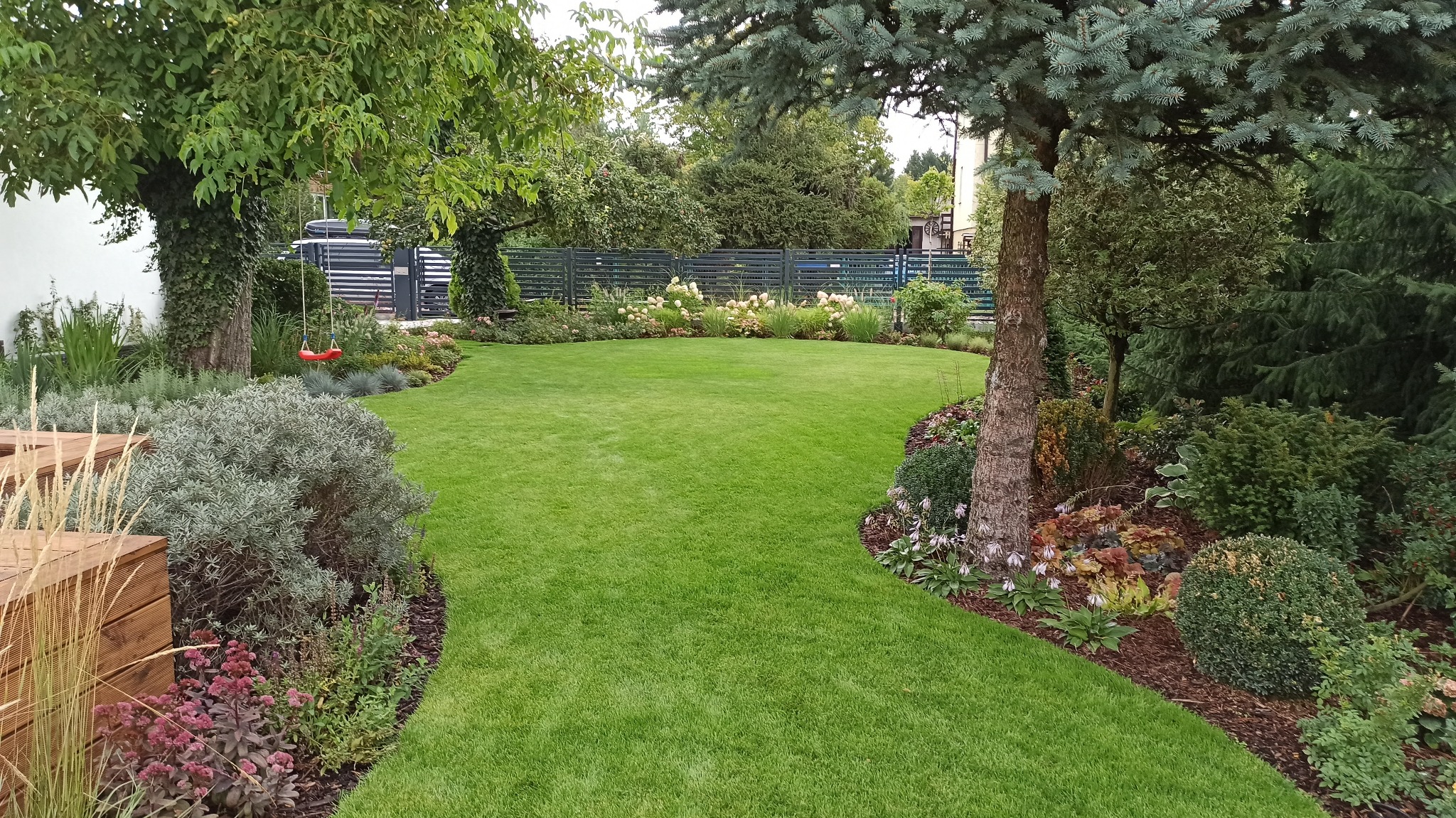 Zadbany trawnik z nasadzeniami i huśtawką w ogrodzie w Nysie, widok na zielony, wypielęgnowany teren z różnorodną roślinnością.