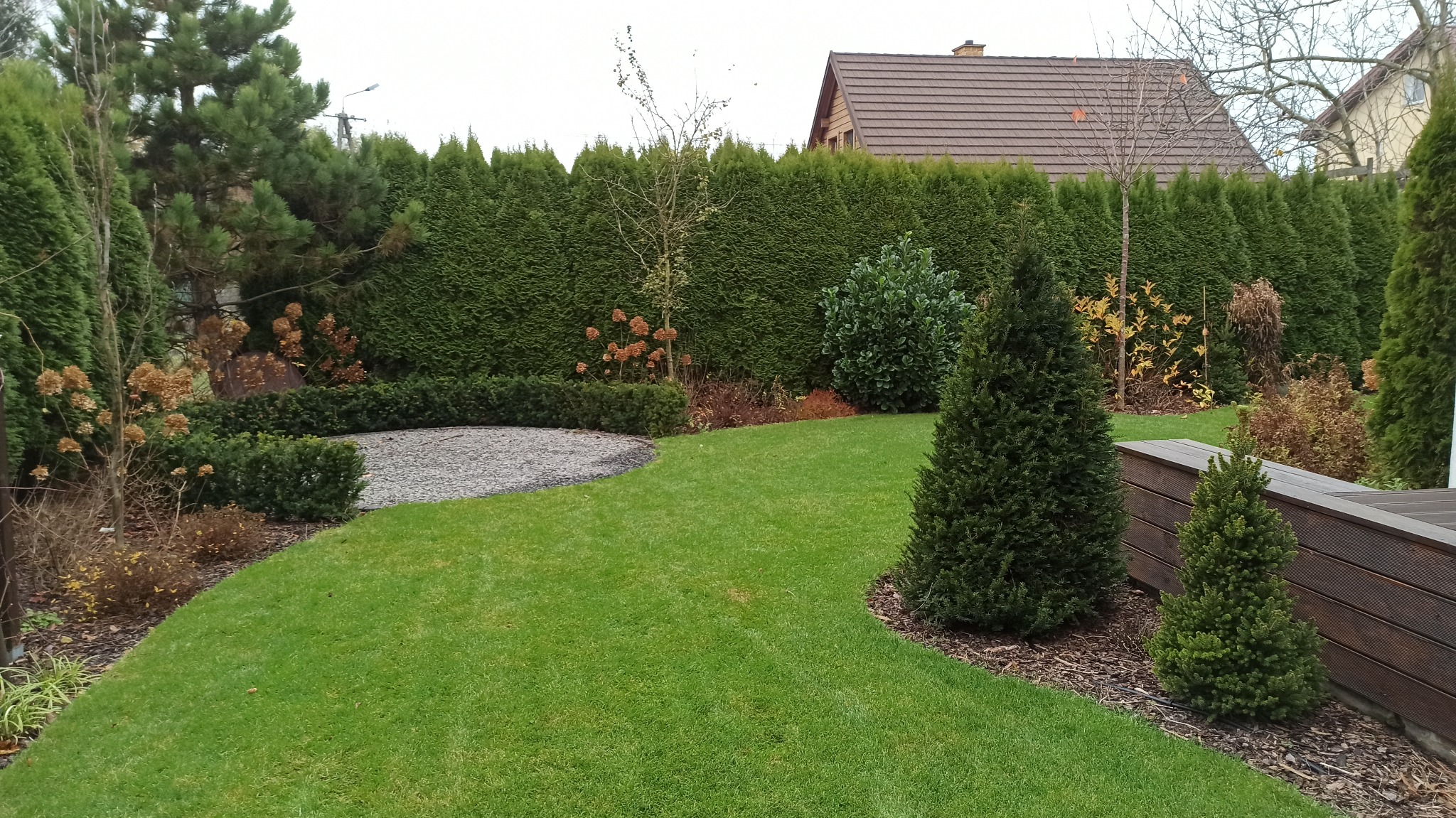 Zadbany ogród z równo przystrzyżonym trawnikiem, szpalerem tuj, żwirowym obszarem i drewnianym obrzeżem rabaty, widok z góry na dom z brązowym dachem.