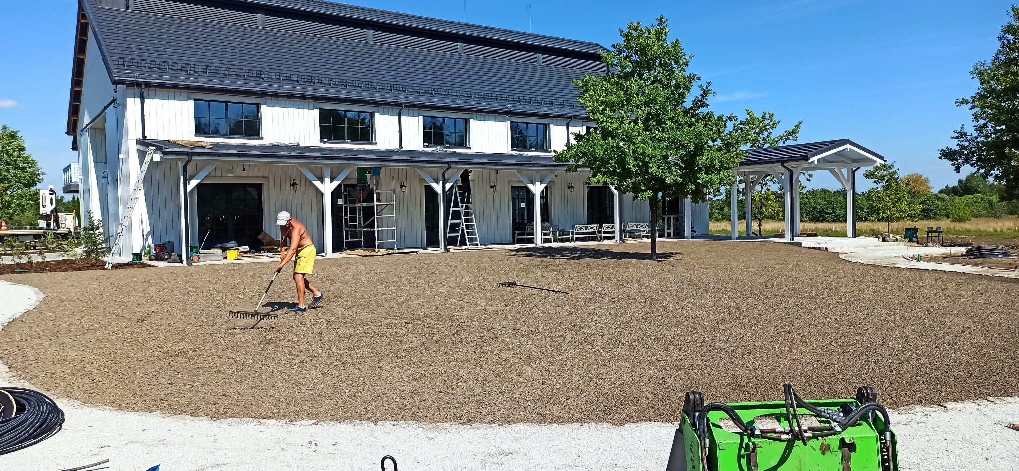Przygotowanie terenu pod trawnik: mężczyzna w żółtych spodenkach wyrównuje grabiami ziemię na rozległym placu przed nowoczesnym, dwukondygnacyjnym budynkiem z białymi ścianami i ciemnym dachem,...