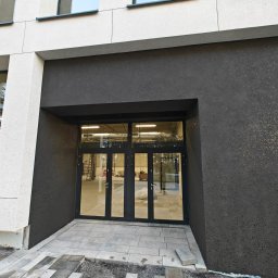 BUDOWNICTWO SERHII SHTYK - Nowoczesna elewacja budynku z ciemnoszarym tynkiem strukturalnym i jasnymi panelami. Wejście z przeszklonymi drzwiami i szarą kostką brukową.