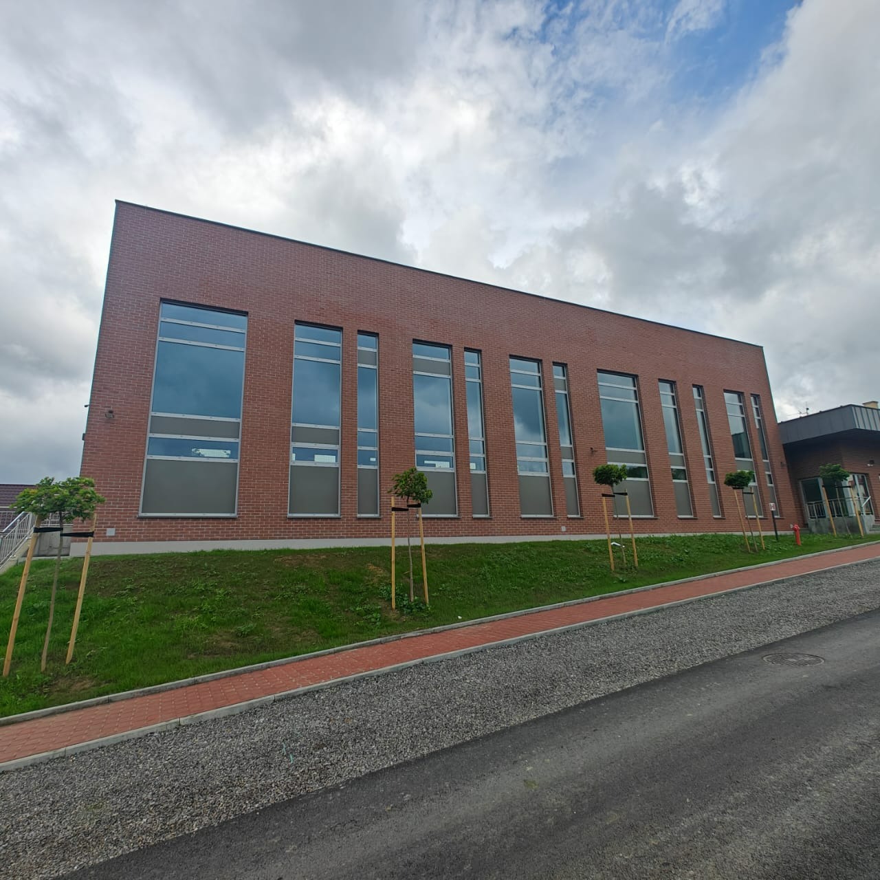 Nowoczesny budynek z ceglaną elewacją i dużymi oknami, otoczony zielenią i chodnikiem z kostki. Widok z dołu, pochmurne niebo w tle.
