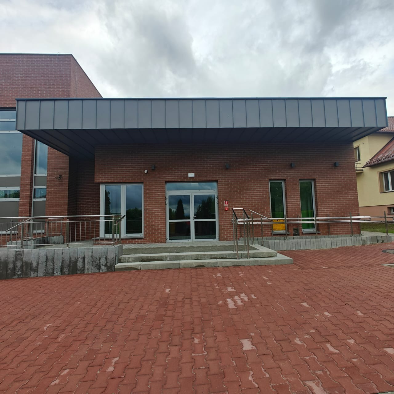 Nowoczesny budynek z ceglaną elewacją, szarym zadaszeniem i przeszklonymi drzwiami wejściowymi. Czerwona kostka brukowa na podjeździe. Pochmurne niebo w tle.