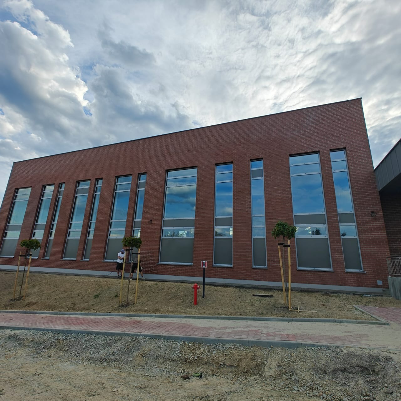 Nowoczesny budynek z cegły klinkierowej z wysokimi oknami, w których odbija się niebo. Widoczny fragment chodnika i trawnika z młodymi drzewkami.