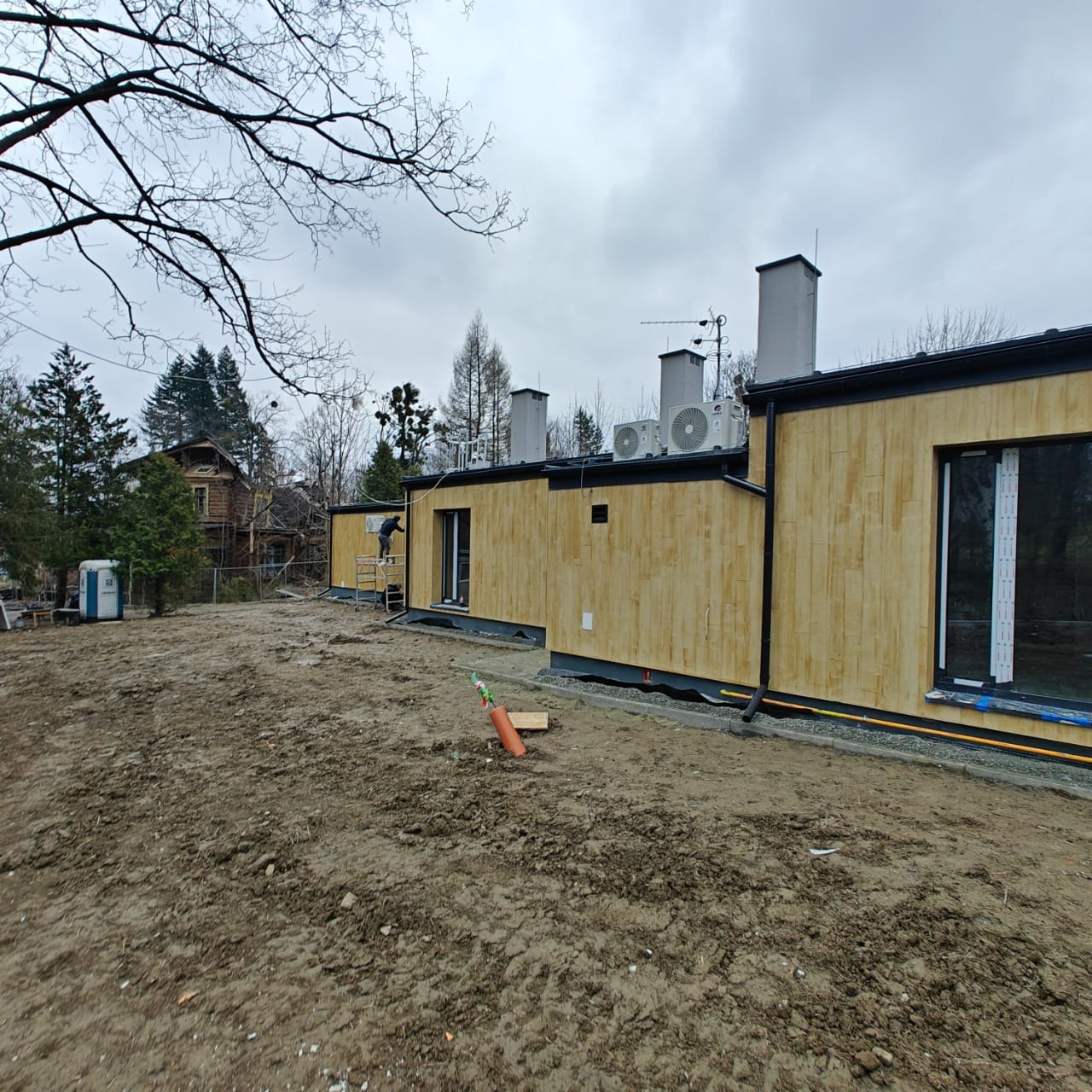 Budynek z drewnianą elewacją w trakcie montażu, widoczny pracownik na rusztowaniu, klimatyzatory na dachu i przygotowany teren wokół.