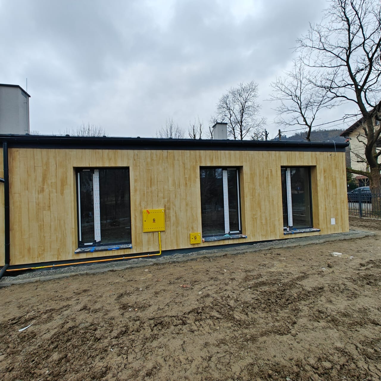 Dom z elewacją imitującą drewno, z trzema oknami i żółtą skrzynką gazową. Widoczne rury gazowe. Tło: pochmurne niebo i drzewa bez liści.