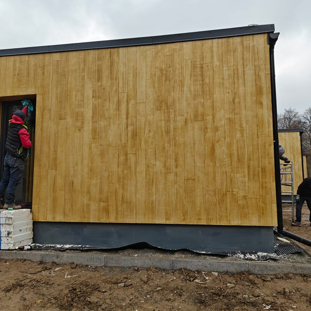 Pracownik czyści elewację budynku o drewnopodobnej fakturze, stojąc na styropianowych blokach. Widoczne detale wykończenia i fragment drabiny z innym pracownikiem w tle.