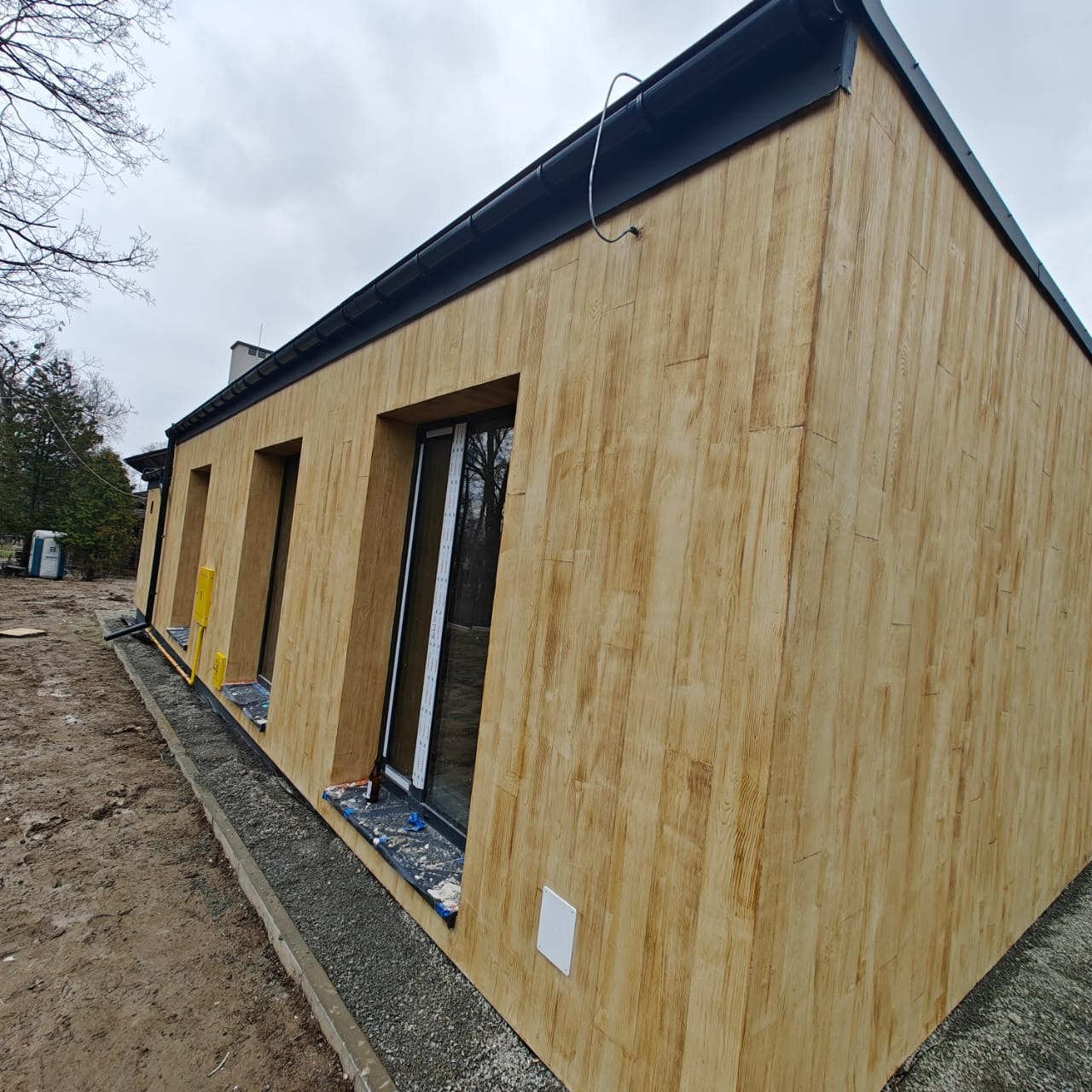 Dom jednorodzinny z elewacją imitującą drewno, widok z boku, czarne rynny, szare niebo, teren wokół domu w trakcie prac wykończeniowych.