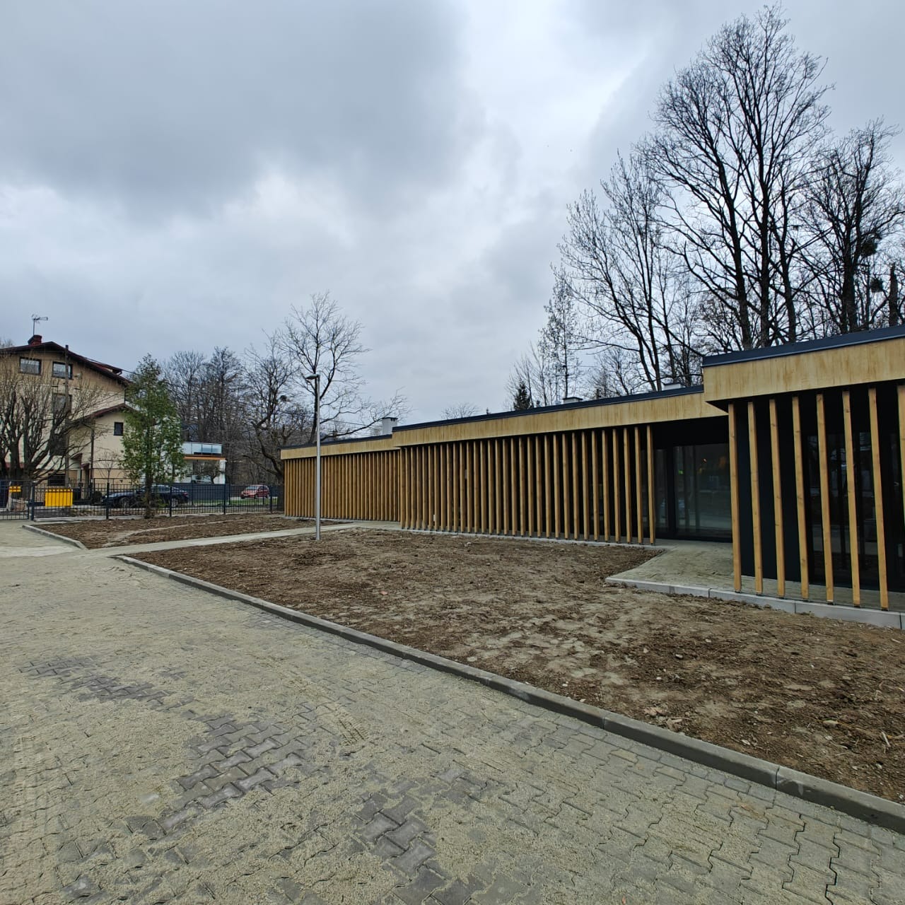 Nowoczesny budynek z drewnianą elewacją w pionowe listwy, widok z zewnątrz, kostka brukowa i fragment budynku mieszkalnego w tle.