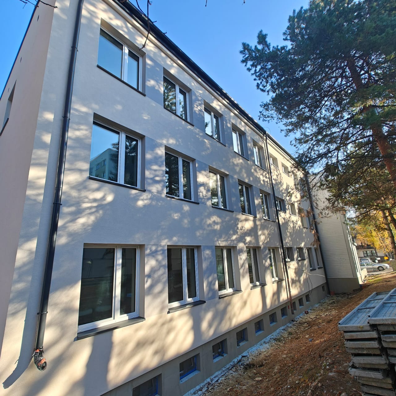 Odnowiona elewacja budynku wielorodzinnego w odcieniach szarości, z widocznymi oknami i elementami wykończeniowymi. Cień drzew na fasadzie, błękitne niebo w tle.