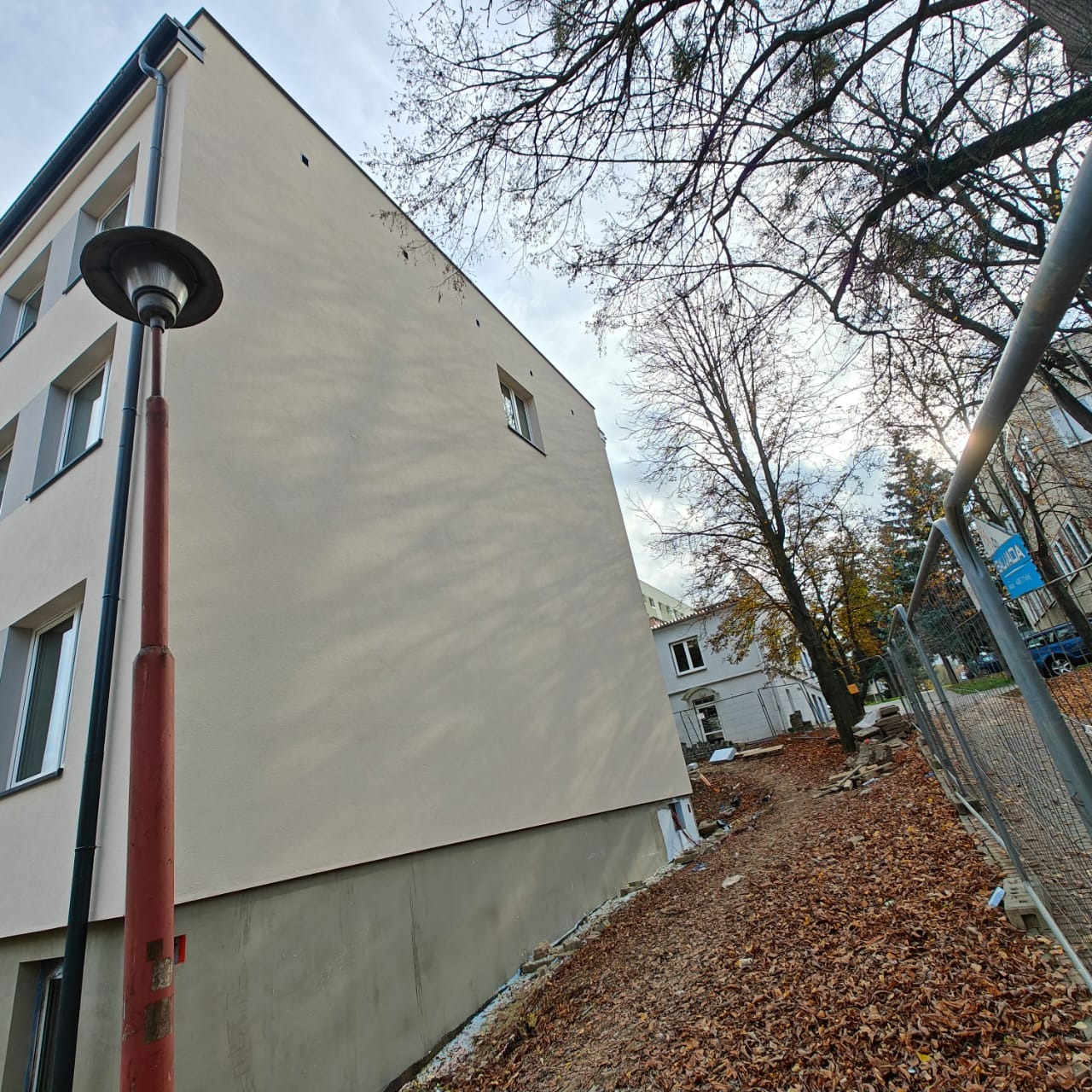 Jasna elewacja budynku wielorodzinnego po remoncie, widok z dołu, z lampą uliczną i jesiennymi liśćmi. Świeżo otynkowana ściana w neutralnym odcieniu beżu.