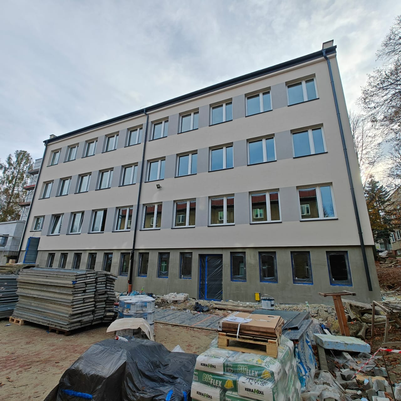 Budynek wielopiętrowy z nową, jasną elewacją w trakcie prac wykończeniowych. Widoczne materiały budowlane i rusztowania. Ujęcie z dołu, pochmurne niebo w tle.