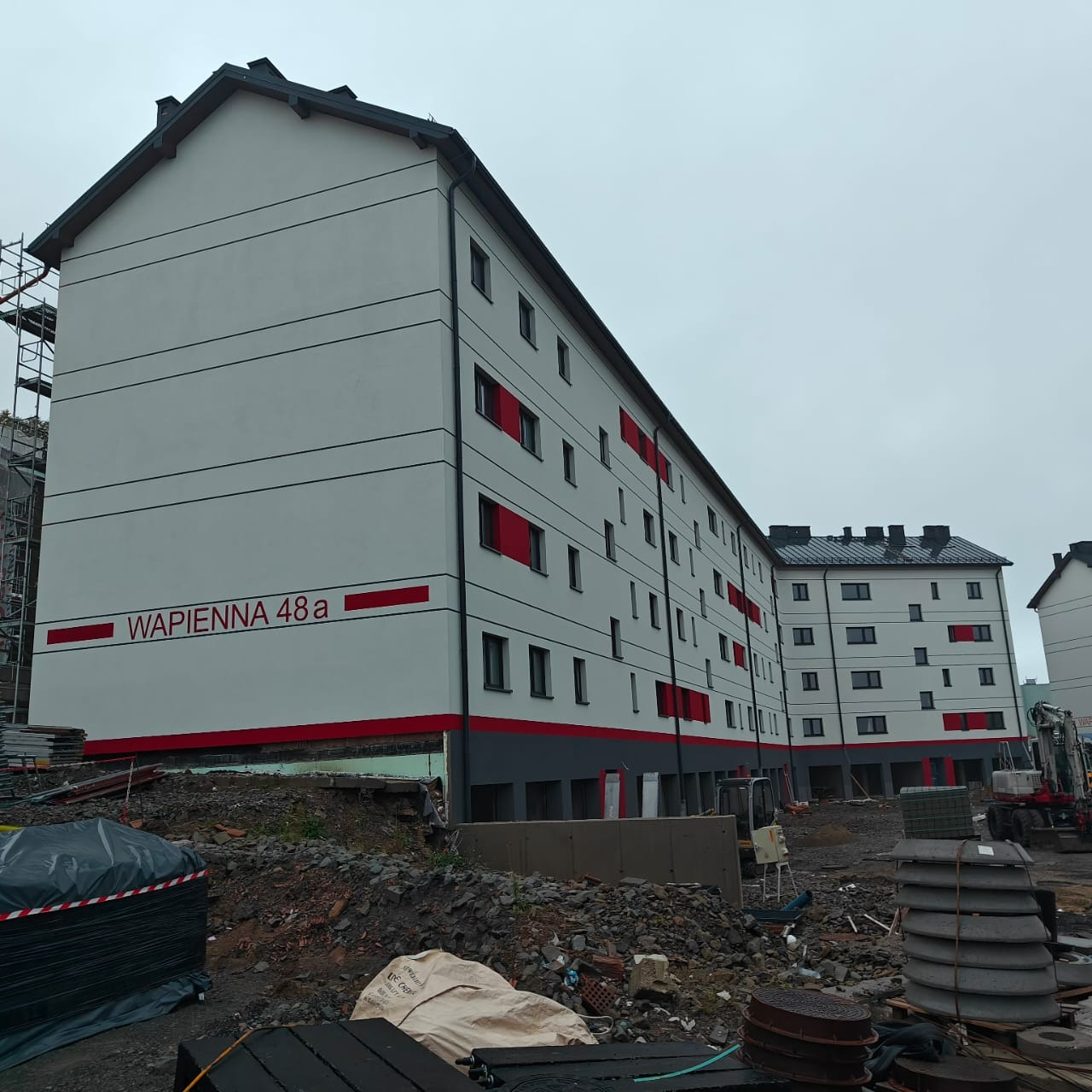 Odnowiona elewacja budynku wielorodzinnego w Pszczynie, z szarymi ścianami, czerwonymi akcentami i czarnymi oknami. Widoczne rusztowania i teren budowy.