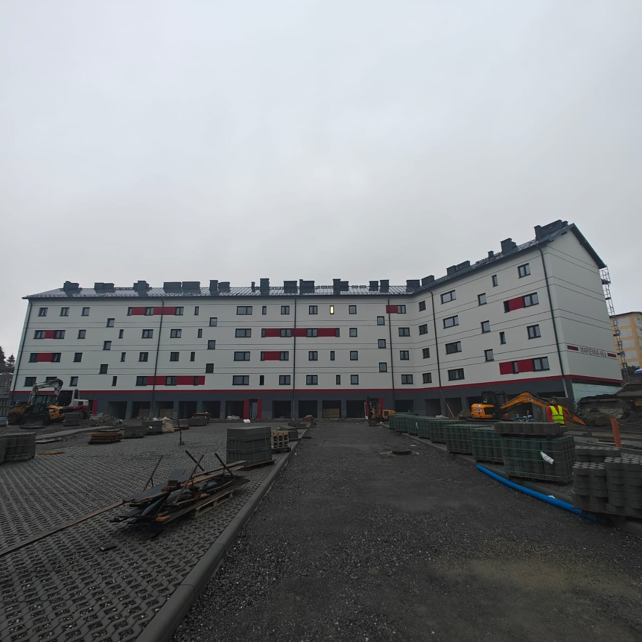 Wielopiętrowy budynek po remoncie elewacji, z białymi ścianami i czerwonymi akcentami, widoczny z placu budowy z kostką brukową i sprzętem budowlanym.