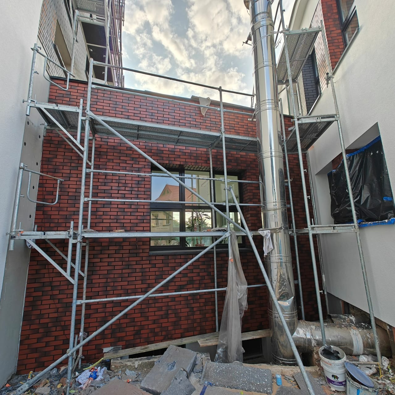 Elewacja z cegły klinkierowej w trakcie montażu, widoczne rusztowania i komin ze stali nierdzewnej. Ujęcie z dołu, fragment nieba.