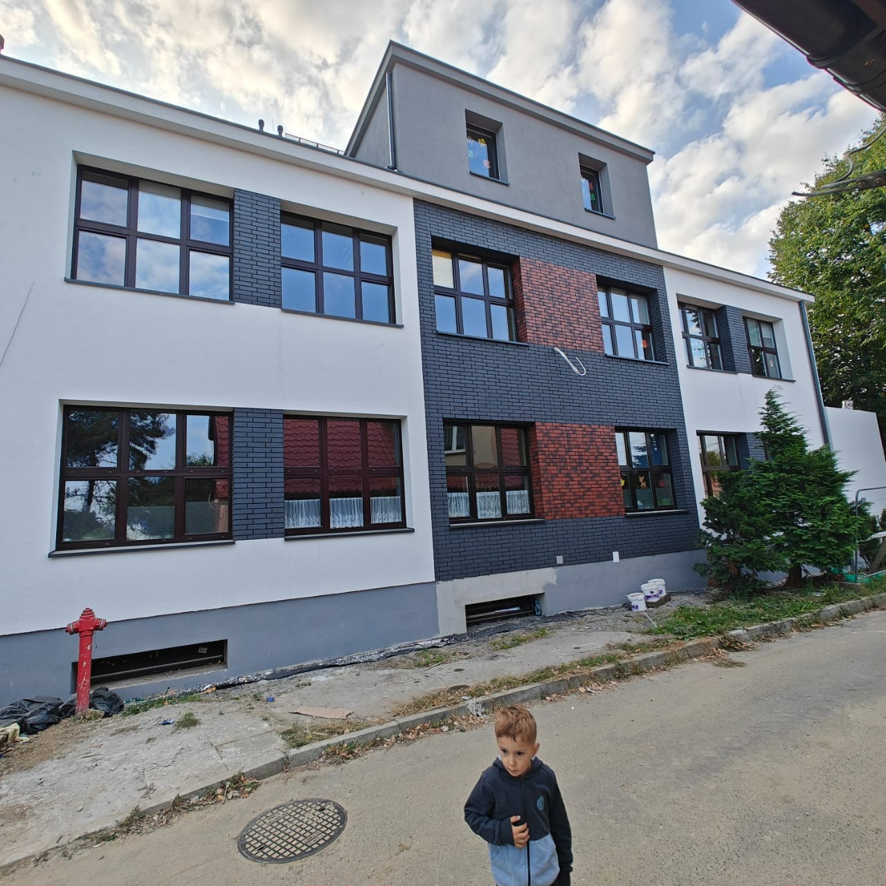 Odnowiona elewacja budynku z elementami cegły klinkierowej i białego tynku, widoczny hydrant i dziecko na pierwszym planie. Świeżo pomalowana fasada.