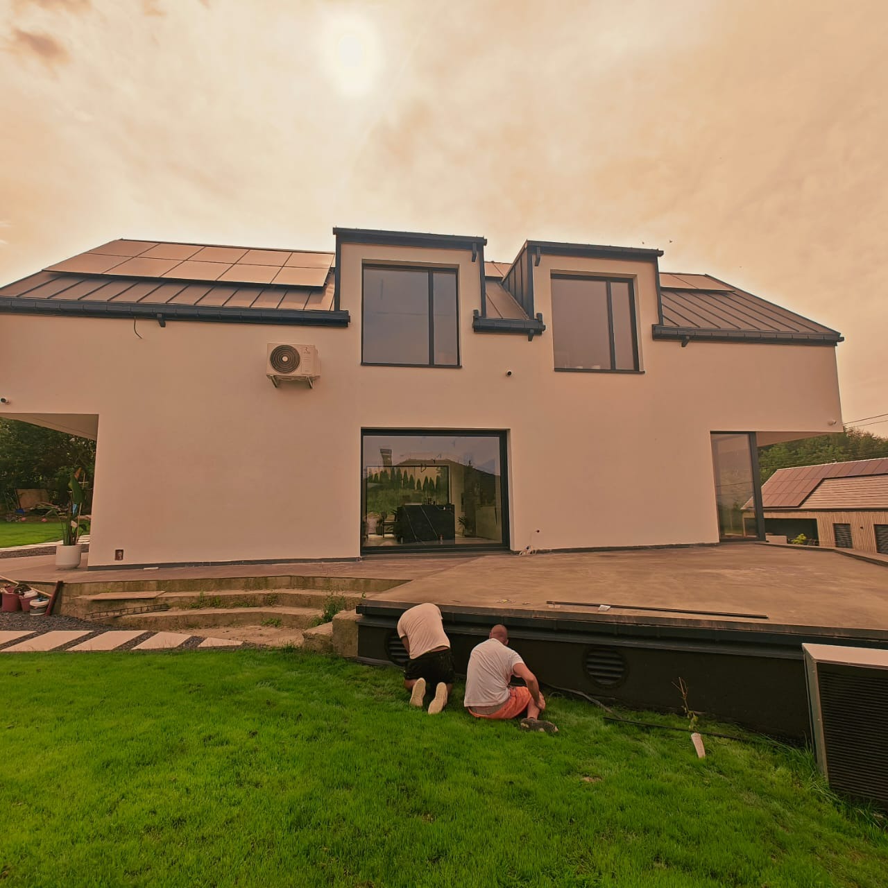 Nowoczesny dom z jasną elewacją i panelami słonecznymi na dachu. Dwóch mężczyzn pracuje na trawniku przed budynkiem, prawdopodobnie przy wykończeniu.