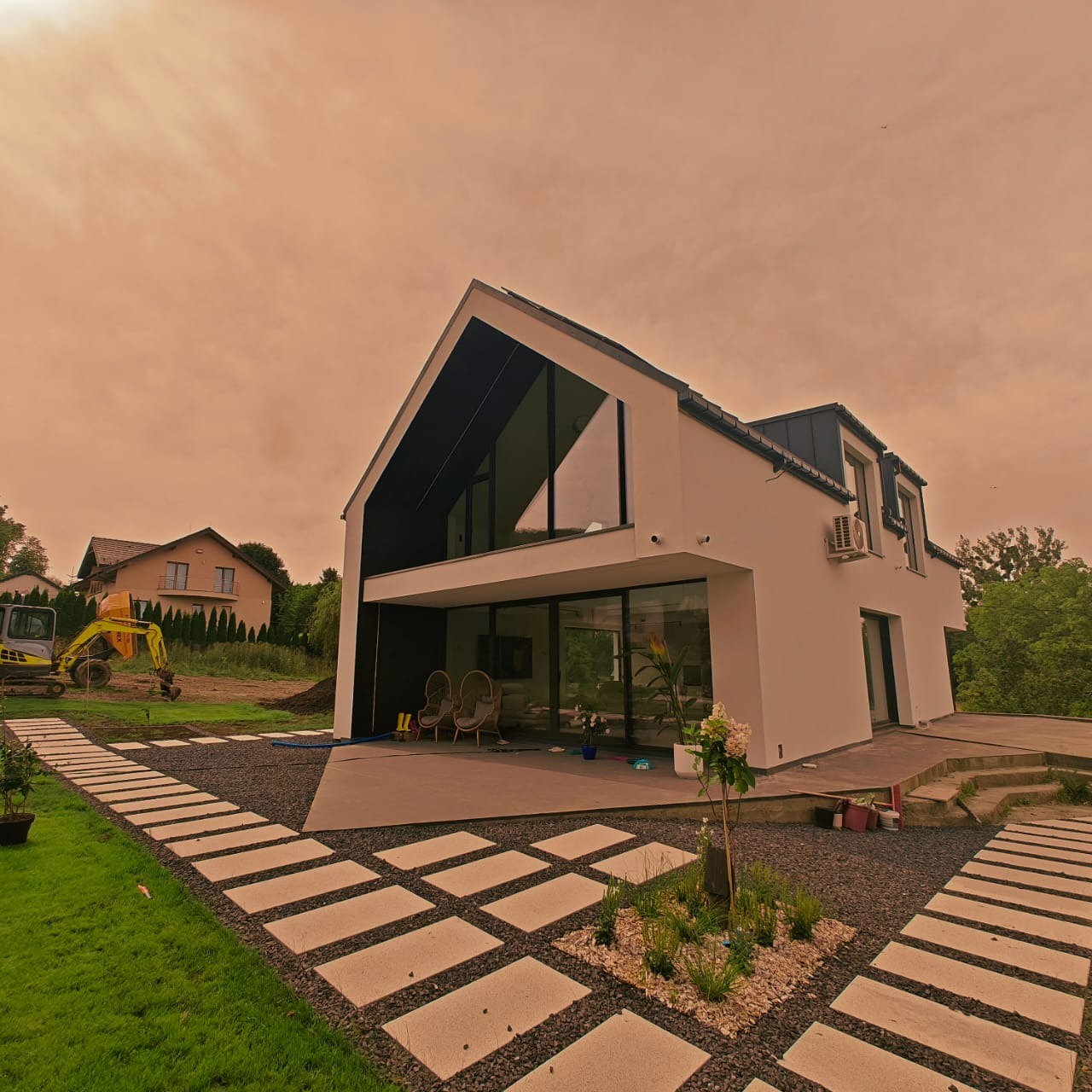 Nowoczesny dom z białą elewacją, dużymi oknami i minimalistycznym ogrodem. W tle widoczny budynek w trakcie budowy i koparka. Nietypowe, pomarańczowe niebo.
